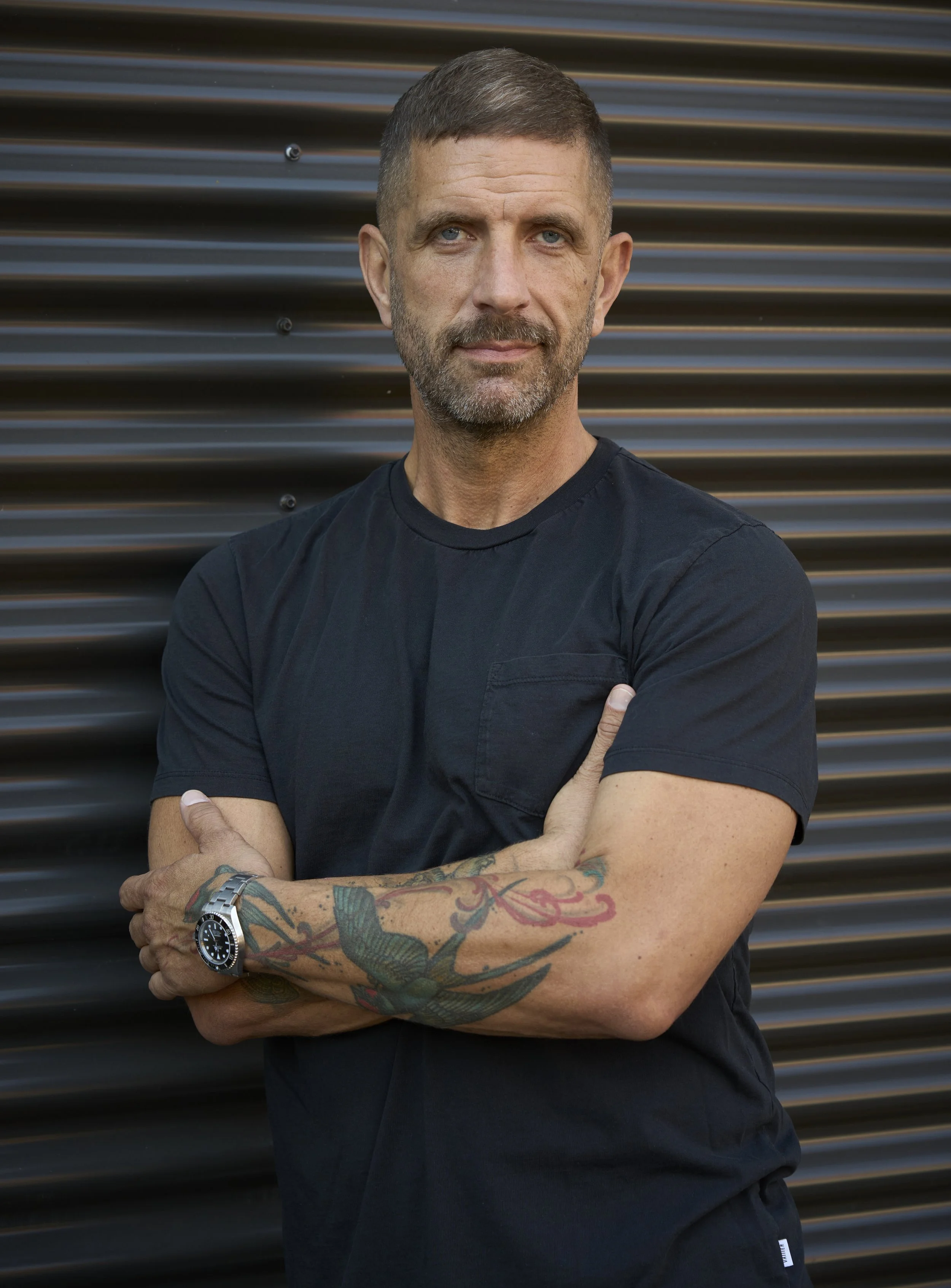 A man with short brown hair and a beard, wearing a black t-shirt, stands with arms crossed in front of a dark, ribbed metal background.