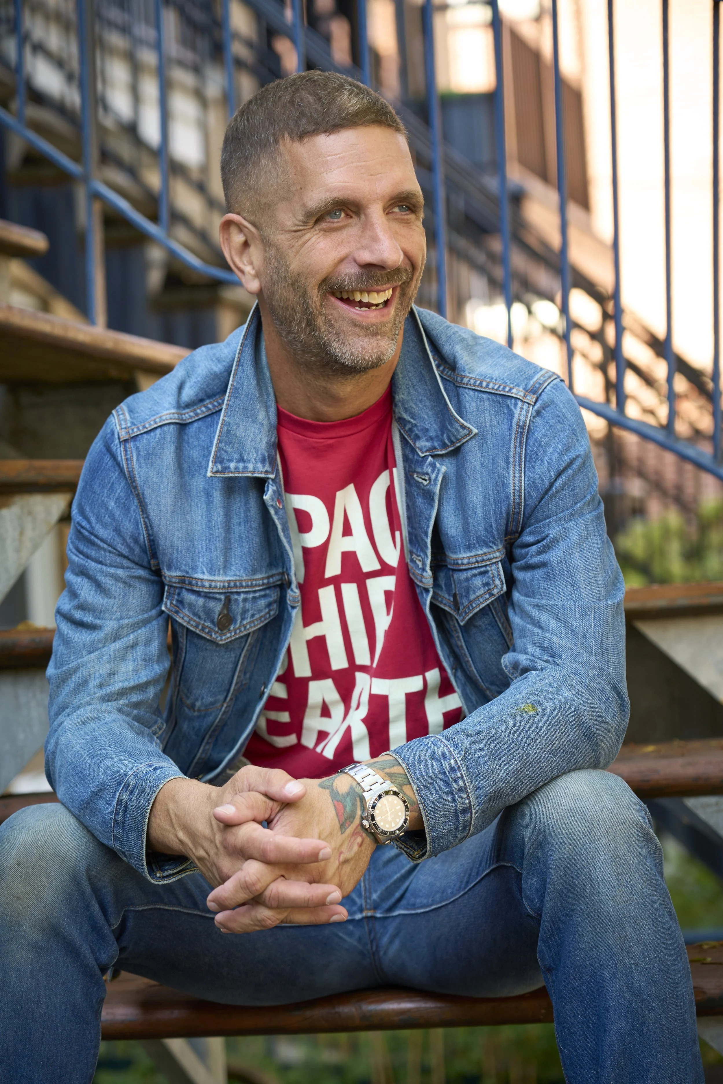 A man sitting on outdoor wooden stairs, smiling, wearing a denim jacket, red T-shirt with white text, and a watch, with blue metal railing in the background.