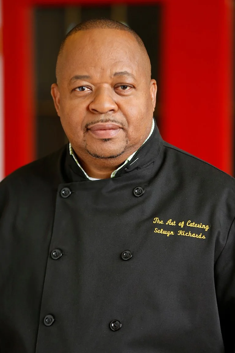 A middle-aged man with short hair wearing a black chef's coat with yellow embroidery reading "The Art of Catering, Selwyn Richards" standing against a blurred red background.