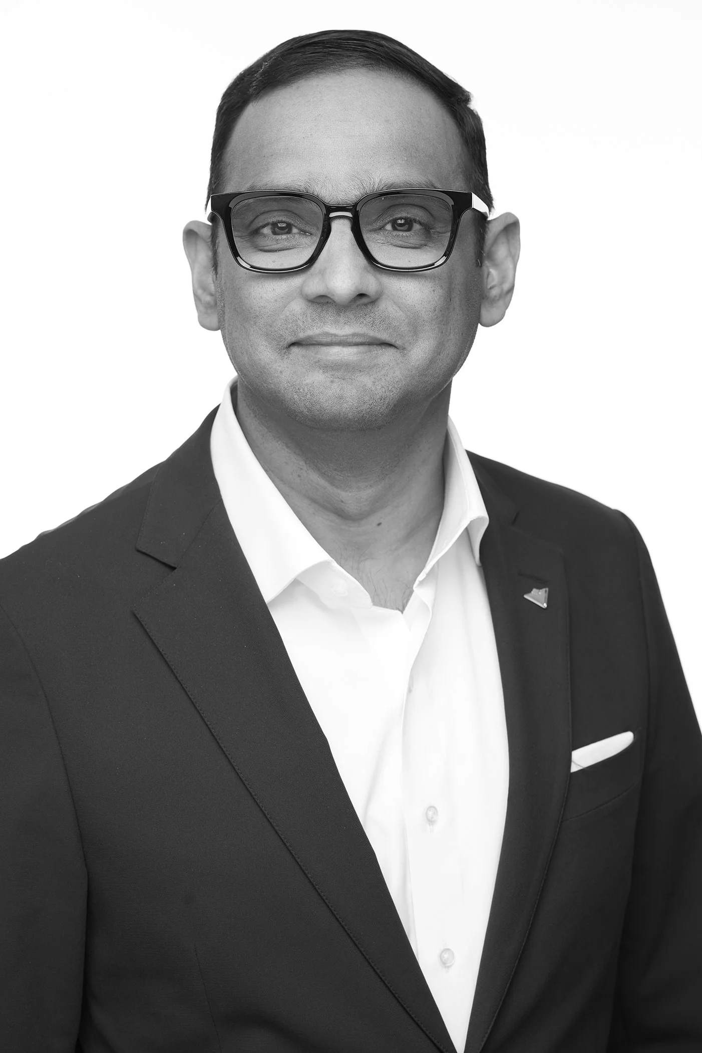 Black and white portrait of a man wearing glasses, a suit, and a white shirt, with a pin on his lapel and a pocket square in his breast pocket, smiling softly.