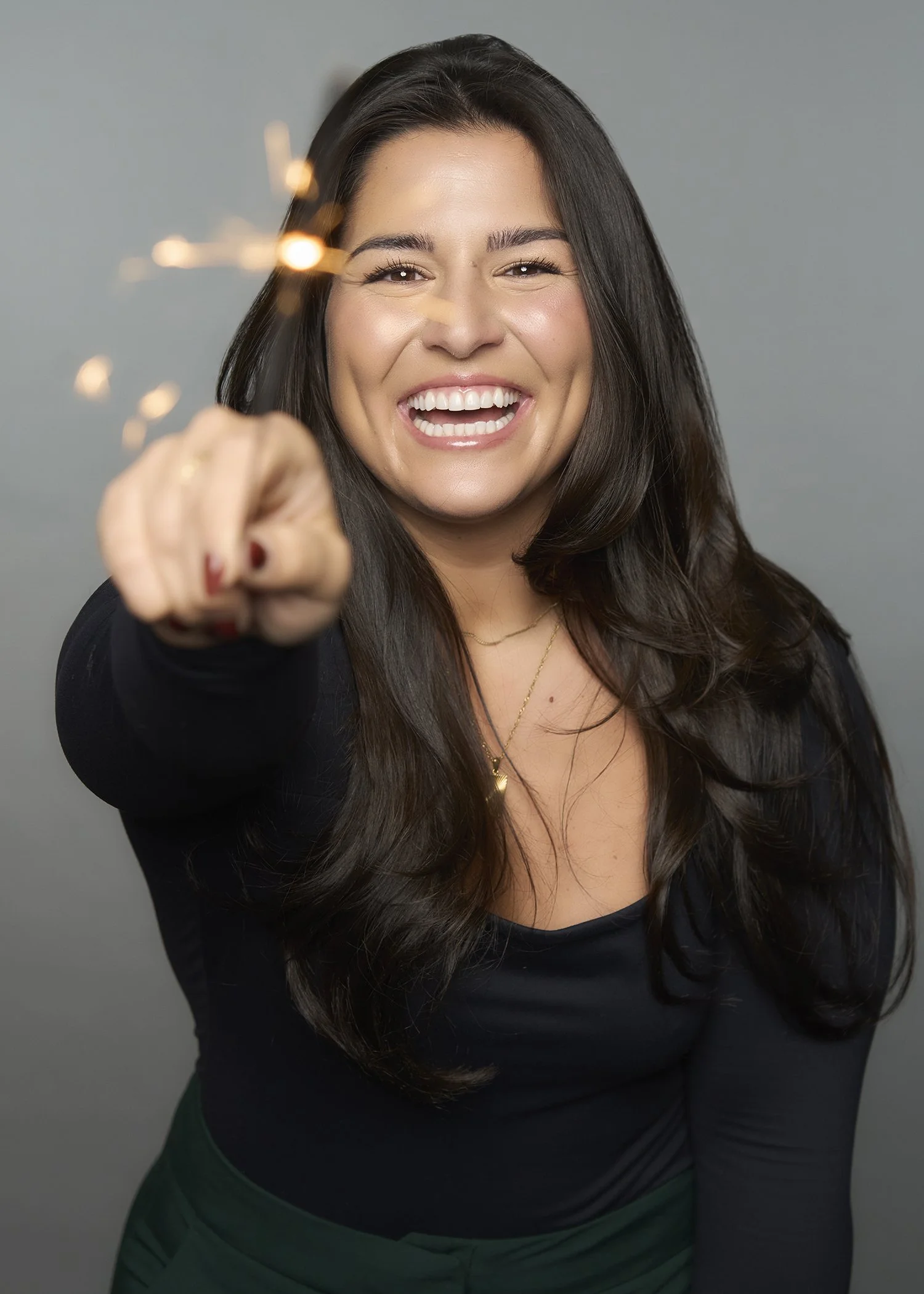 A woman with long dark hair smiling and pointing a sparkler at the camera, wearing a black top and green pants.
