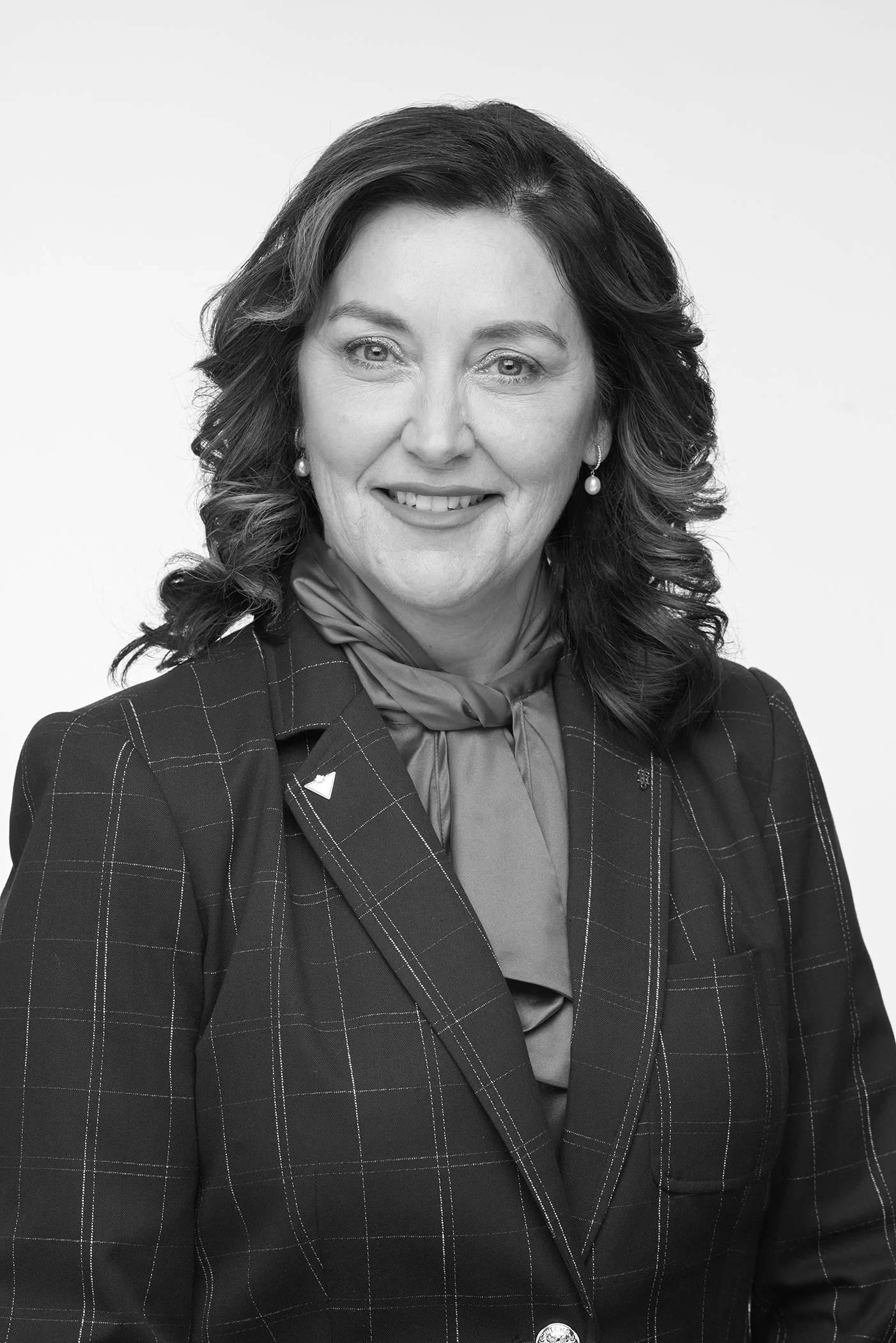 Black and white headshot of a smiling woman with wavy hair, wearing a checkered blazer, a blouse with a bow tie, and pearl earrings.