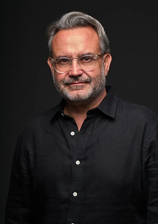 A middle-aged man with gray hair, glasses, and a beard, wearing a black button-up shirt, posing against a dark background.