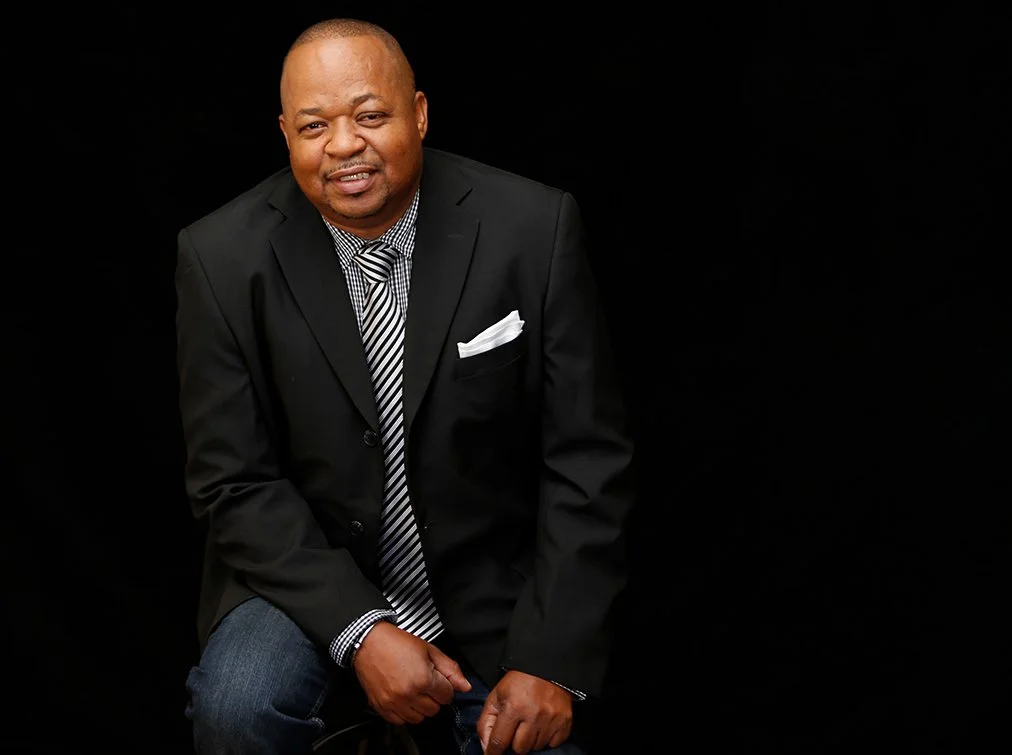 A man in a black suit, checkered shirt, and striped tie posing against a black background.