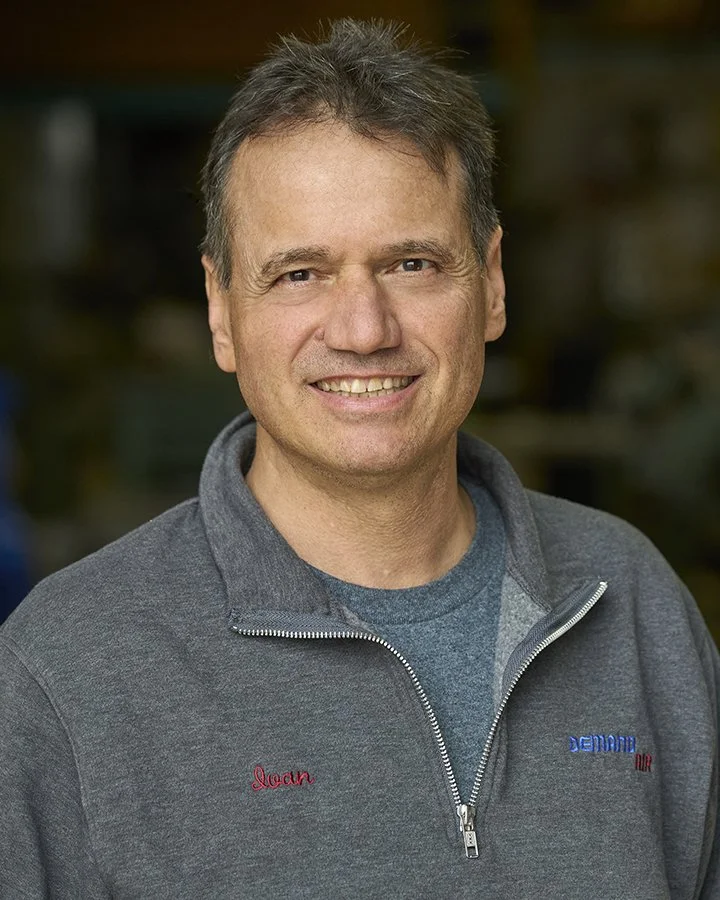 A man smiling, wearing a gray jacket with a zipper and a gray T-shirt underneath. The jacket has the name "Joan" embroidered in red on the left side and the word "Dematic" embroidered in blue on the right side.