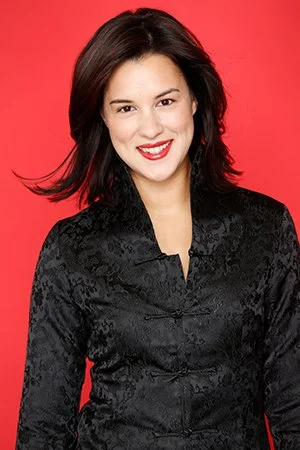 A woman with dark hair smiling, wearing a black jacket, against a red background.