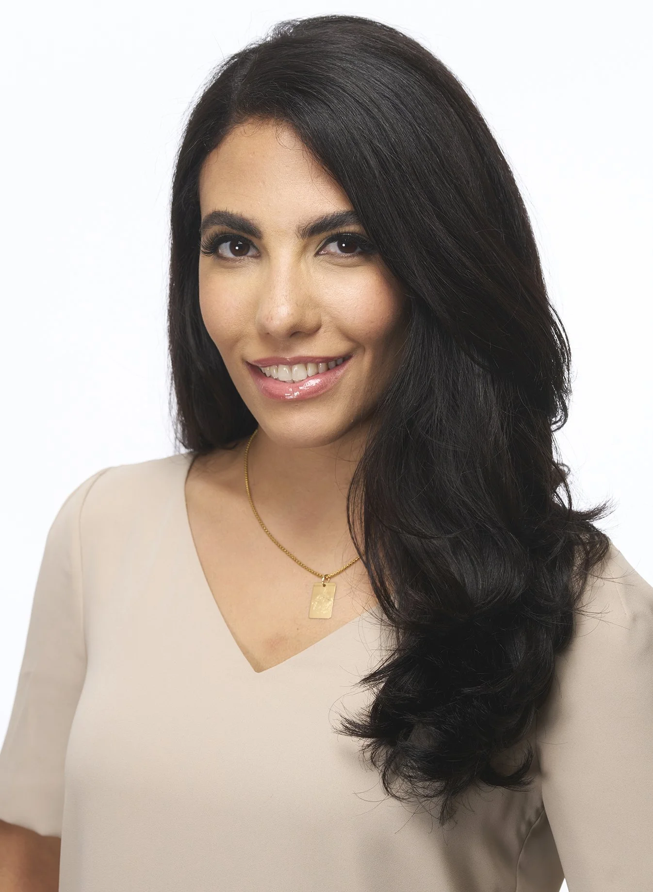 A woman with long dark hair, wearing a beige top and a gold necklace, smiling at the camera against a white background.