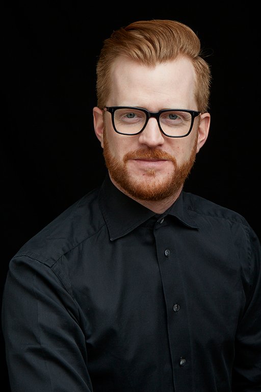 A man with red hair, glasses, and a beard wearing a black shirt against a black background.