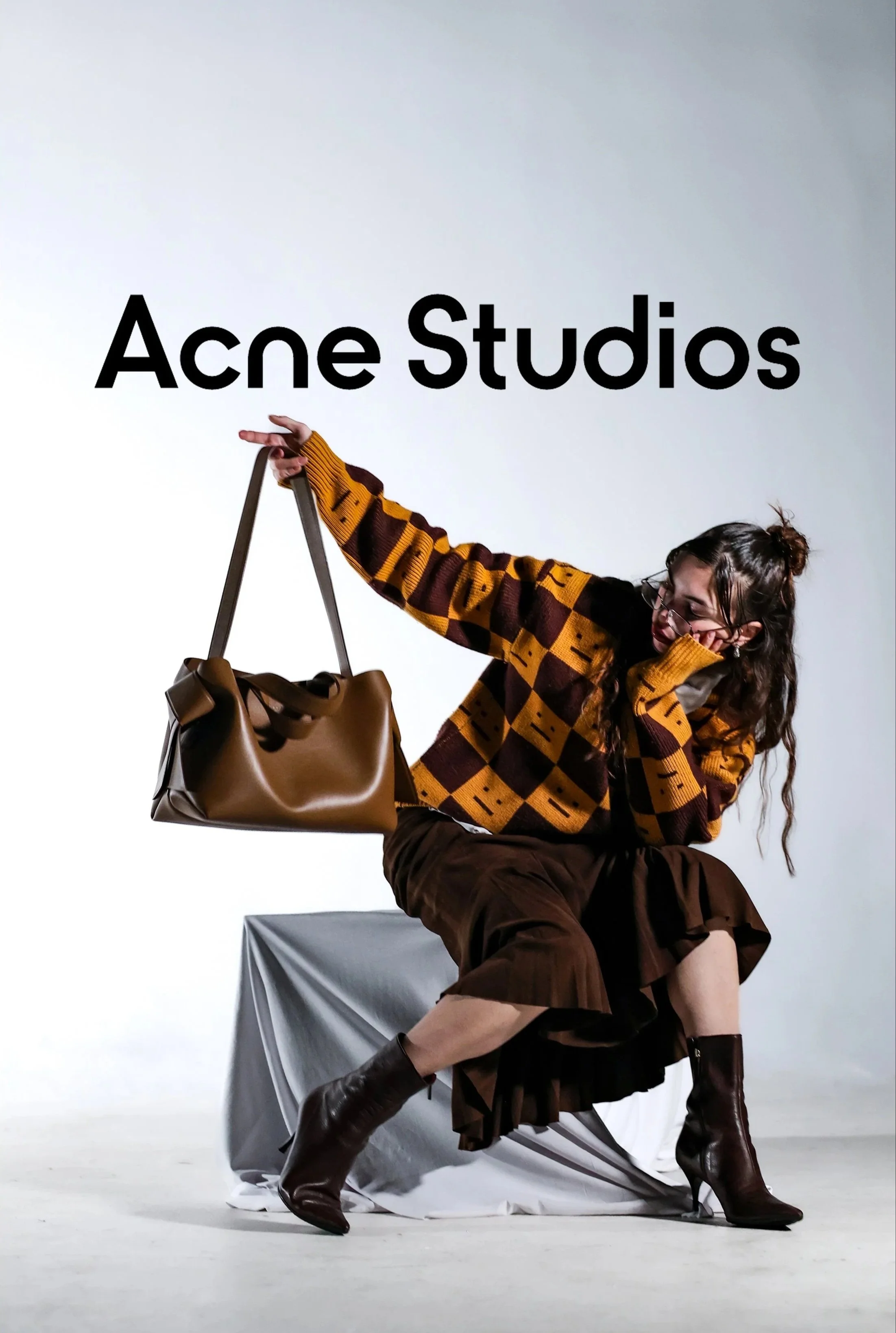 A woman wearing glasses, a checkered sweater, and a brown skirt and boots, sitting on a draped chair and holding a large brown handbag while posing against a plain backdrop with the text "Acne Studios" displayed above.