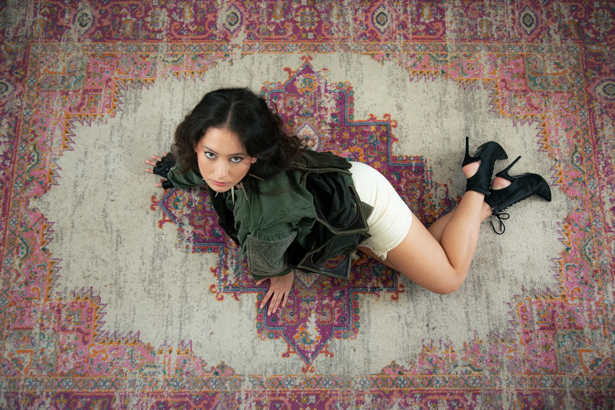 A woman with dark, wavy hair and makeup, wearing a green jacket, cream shorts, and black high-heeled boots, lying on her stomach on a patterned rug and looking up at the camera.