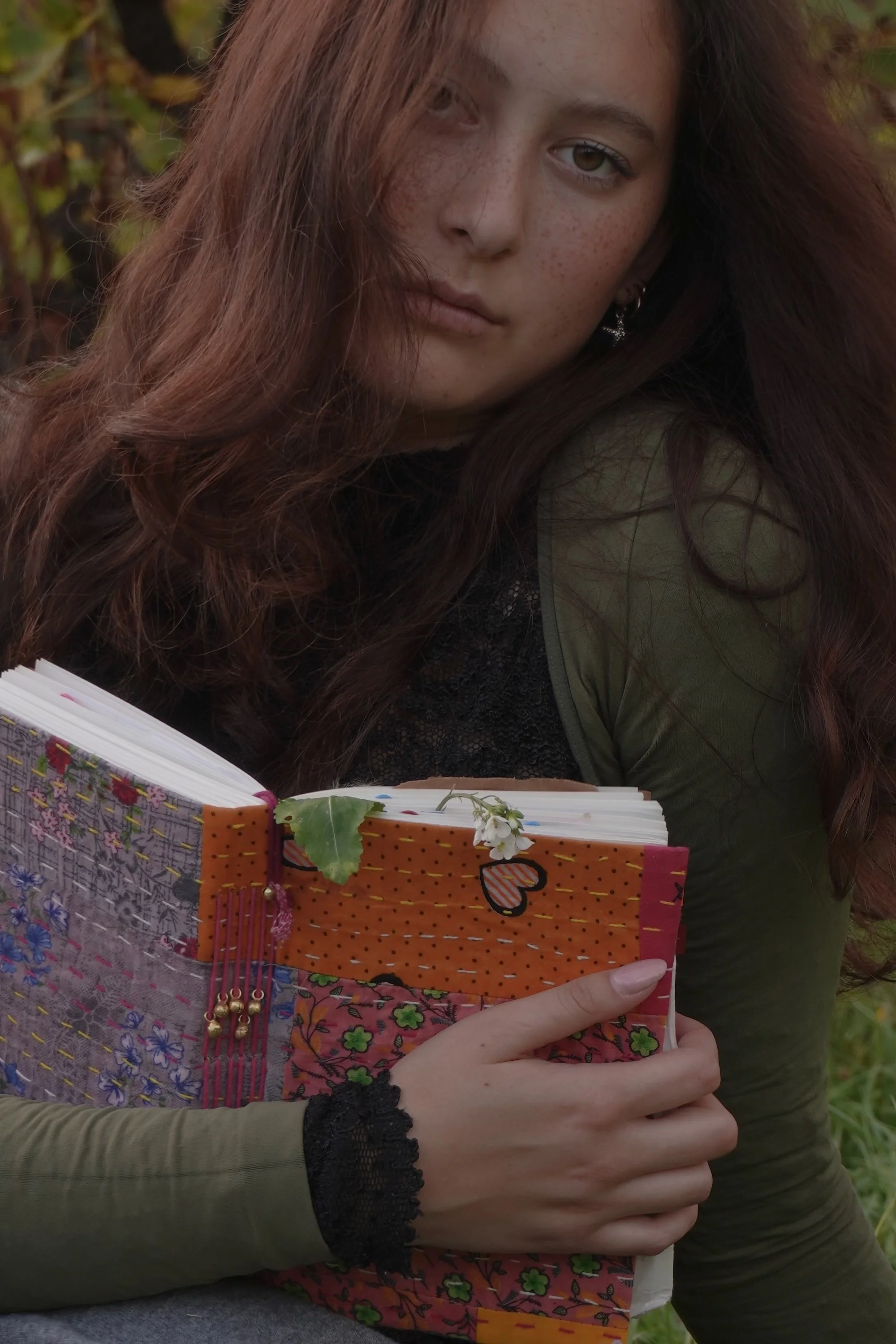 A young woman with long, wavy red hair and freckles, wearing a green jacket, holding a colorful handmade journal with floral and heart decorations, outdoors among foliage.
