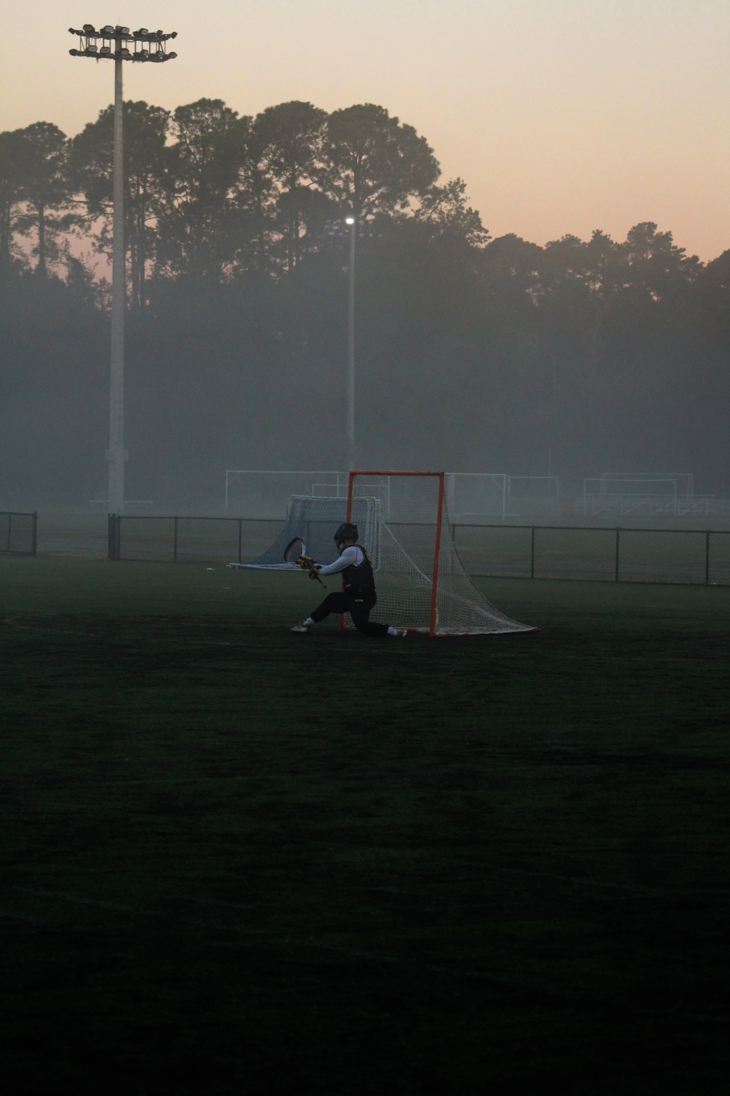 Lacrosse goalkeeper in uniform, kneeling in front of a lacrosse goal, on a field at dusk with fog and trees in the background.
