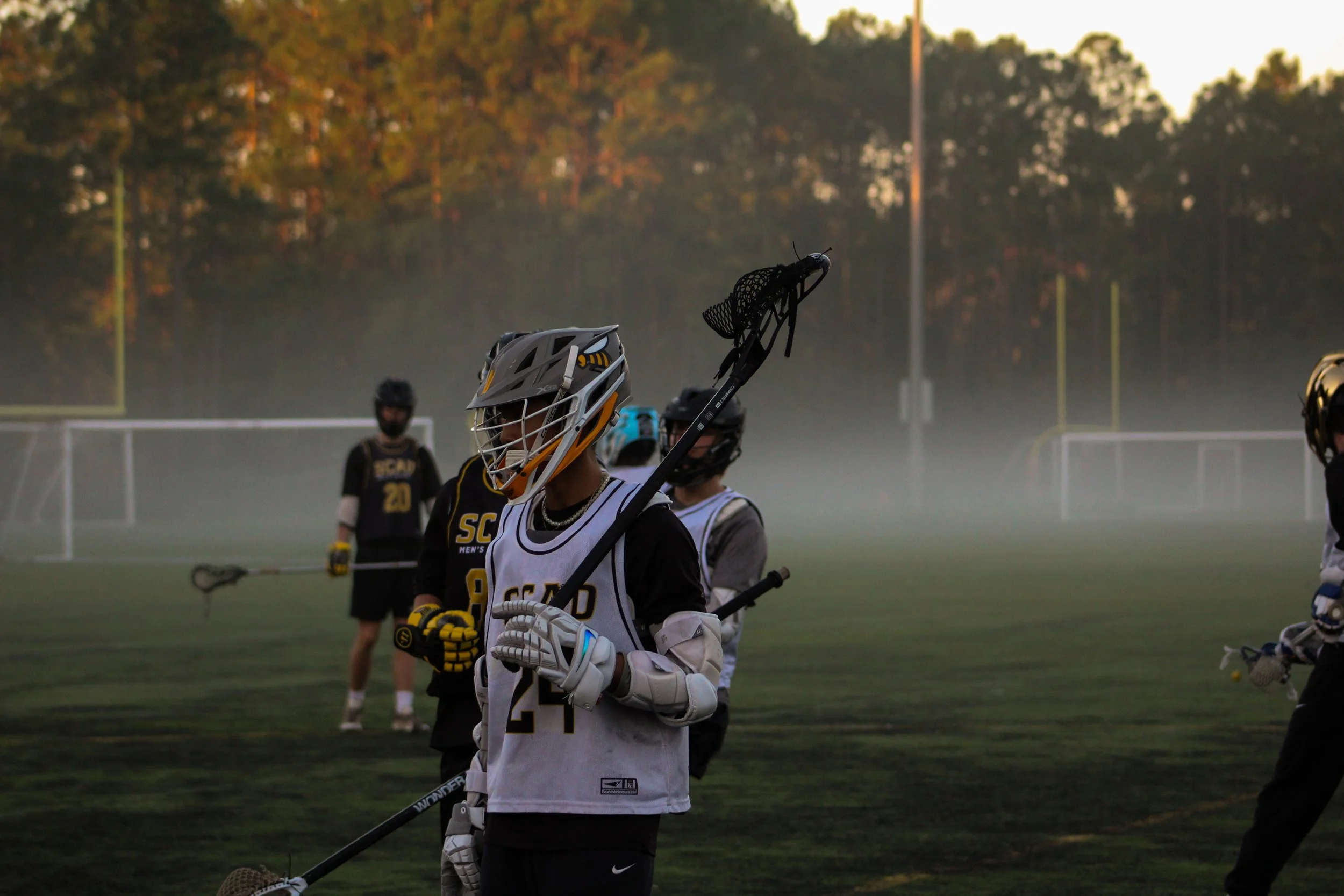 Lacrosse players on a field during a game or practice session at sunrise or sunset with mist in the background.