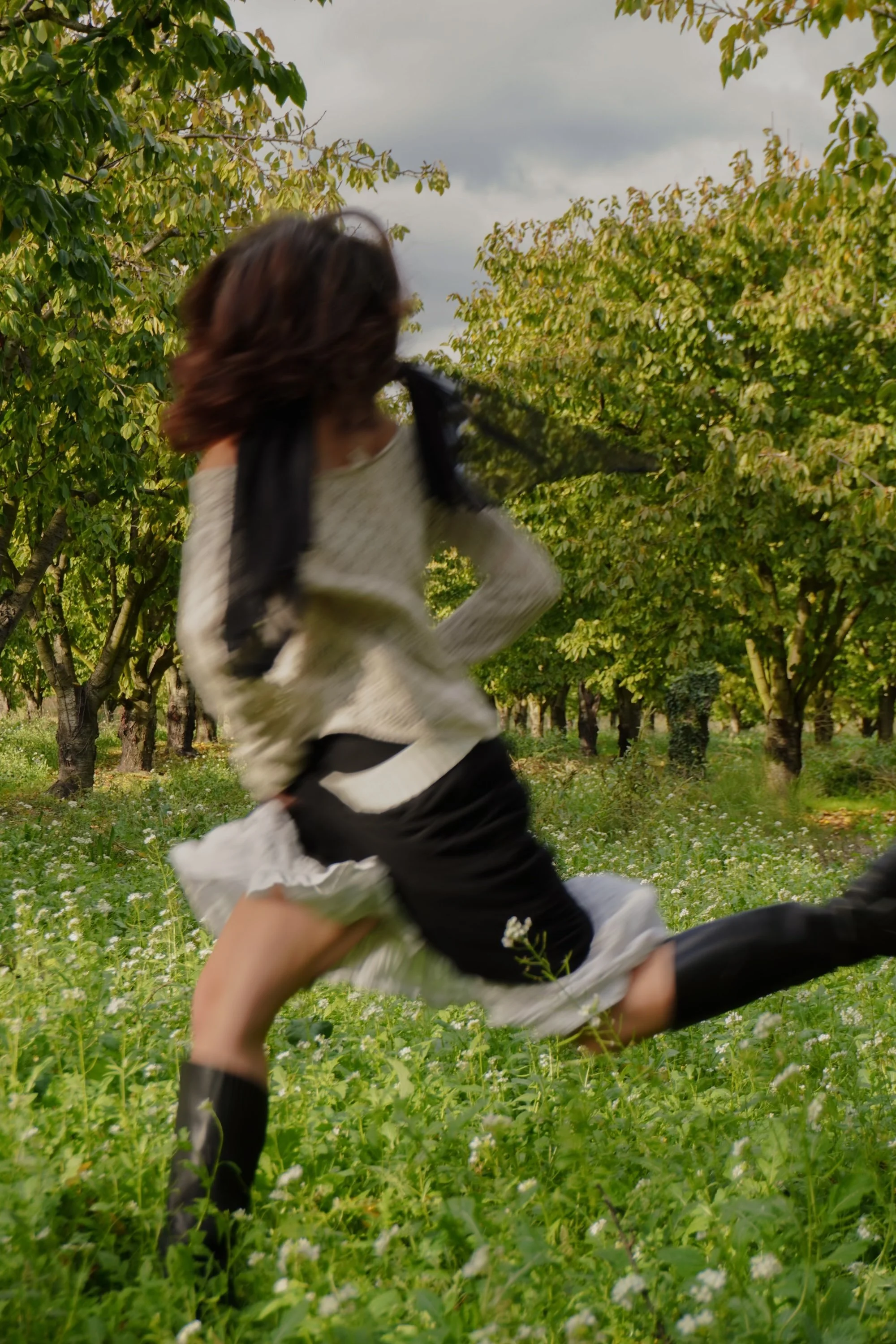 A person running through a green field with trees, wearing a cream sweater, black skirt, and black knee-high boots, holding a plastic bag.