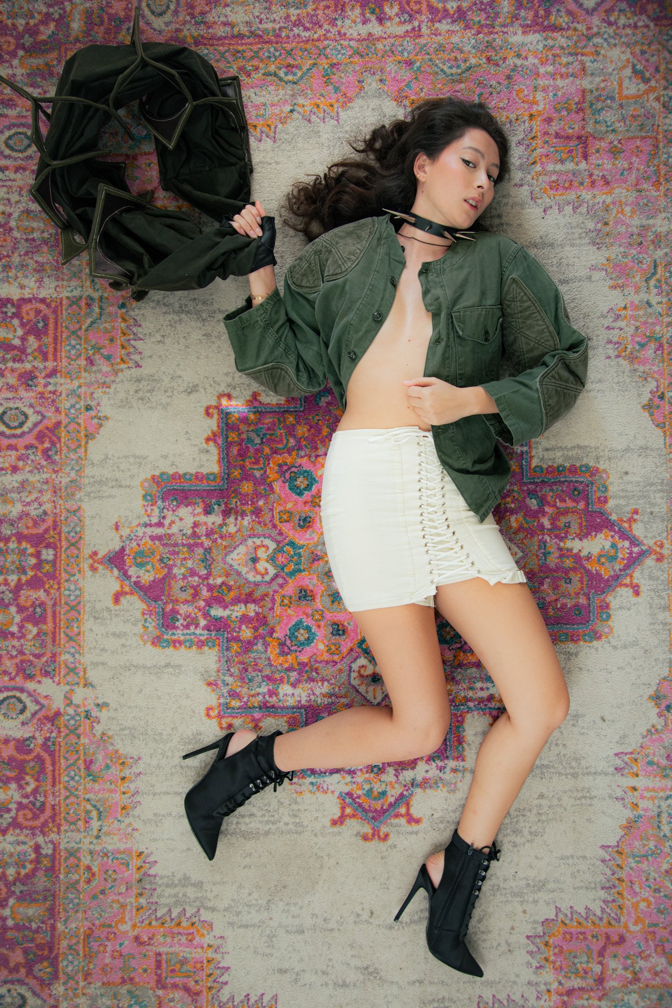 A woman lying on a colorful, patterned rug. She is wearing a green jacket, white lace-up skirt, black high-heeled ankle boots, and a black choker. She has long dark hair and is holding a black jacket.