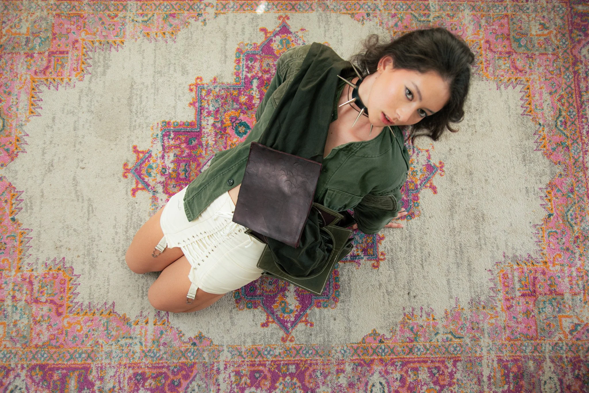 A young woman with dark curly hair, wearing a green jacket and white shorts, kneeling on a decorative multi-colored rug with a vintage pattern, holding black handbags, and wearing a spiked choker necklace, looking up at the camera.