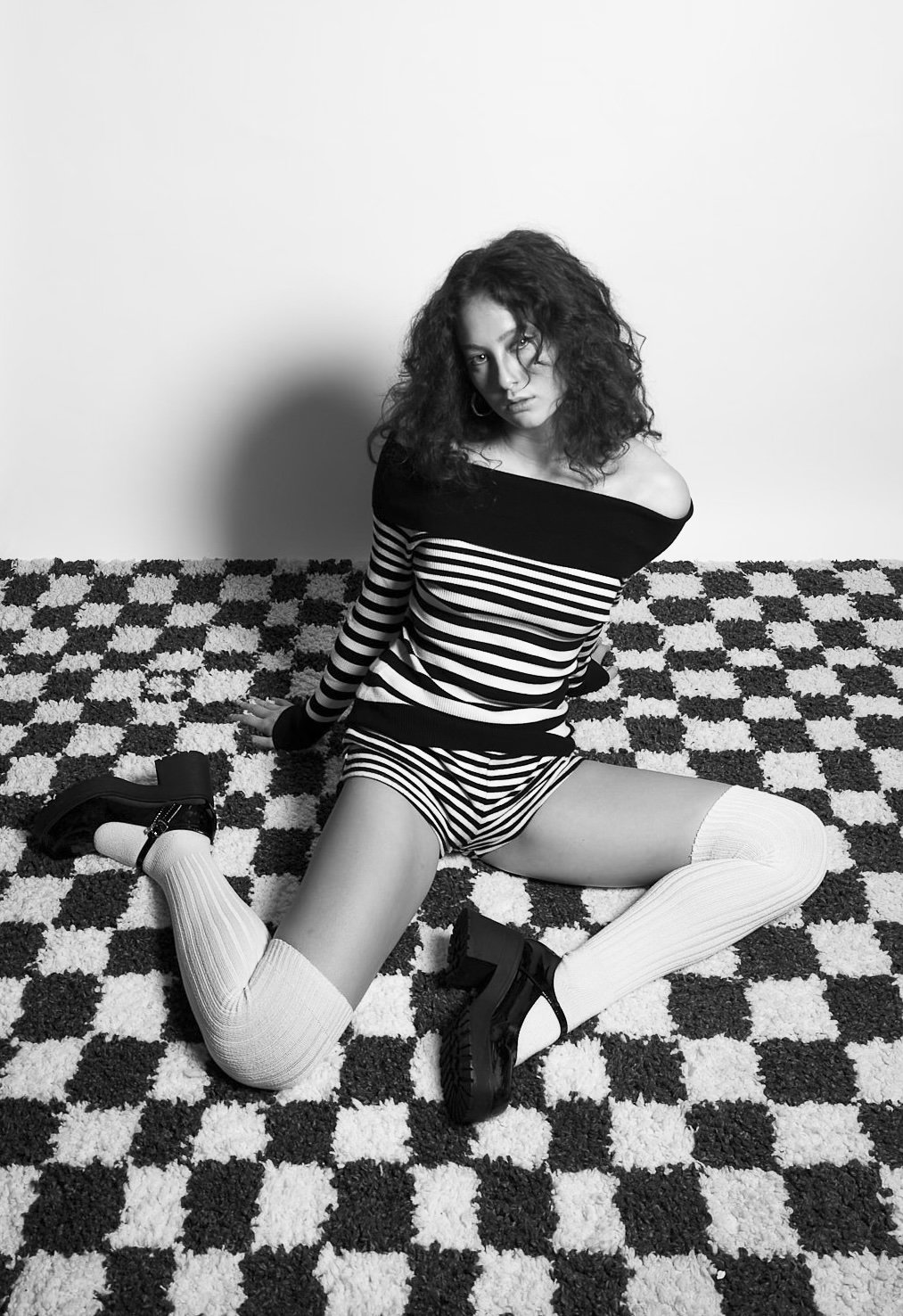 A young woman with curly hair sitting on a checkered carpet against a plain wall, dressed in a striped off-the-shoulder top, striped shorts, and thigh-high socks with black platform shoes, looking at the camera.