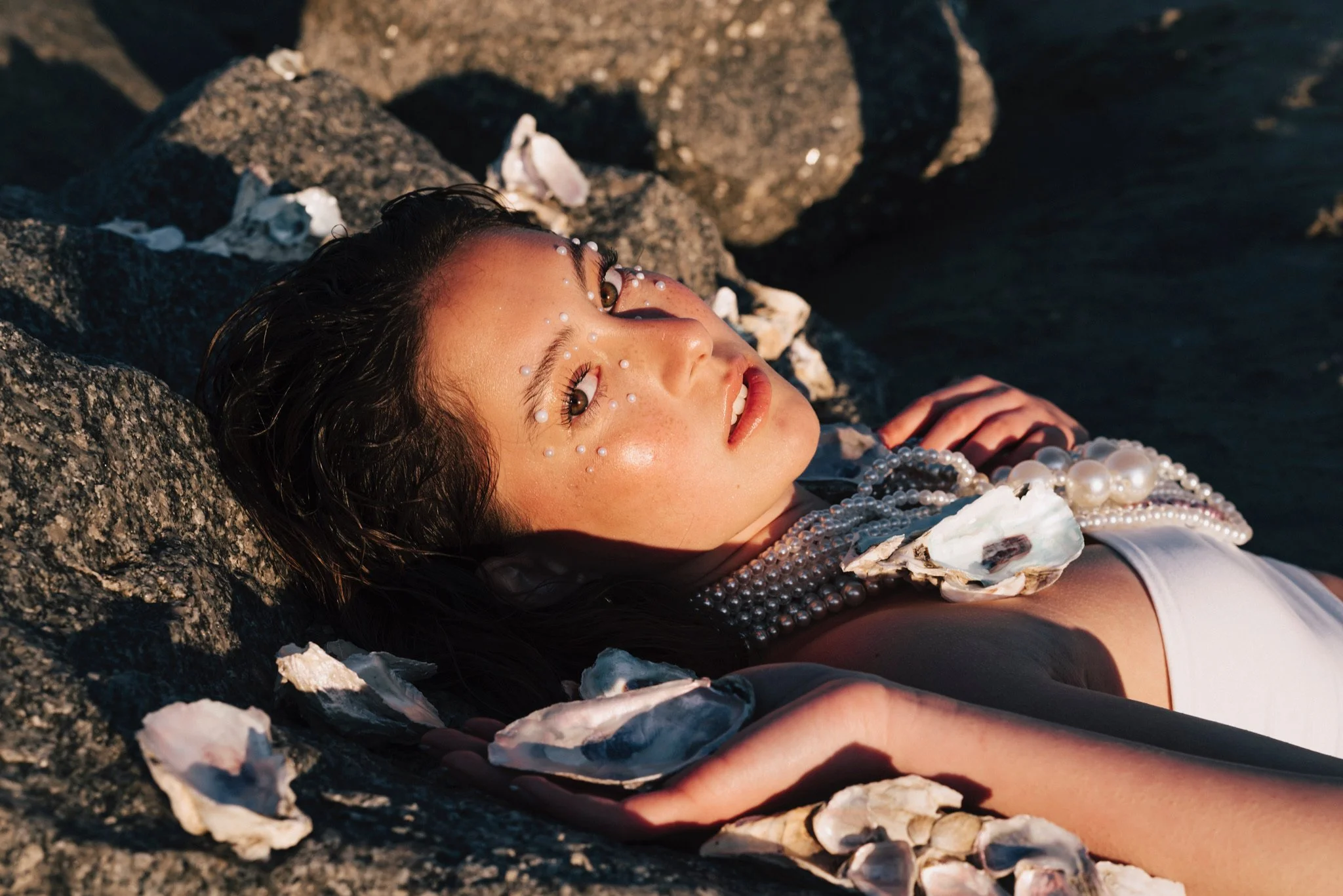 A woman with pearl and shell accessories lying on rocks by the water at sunset
