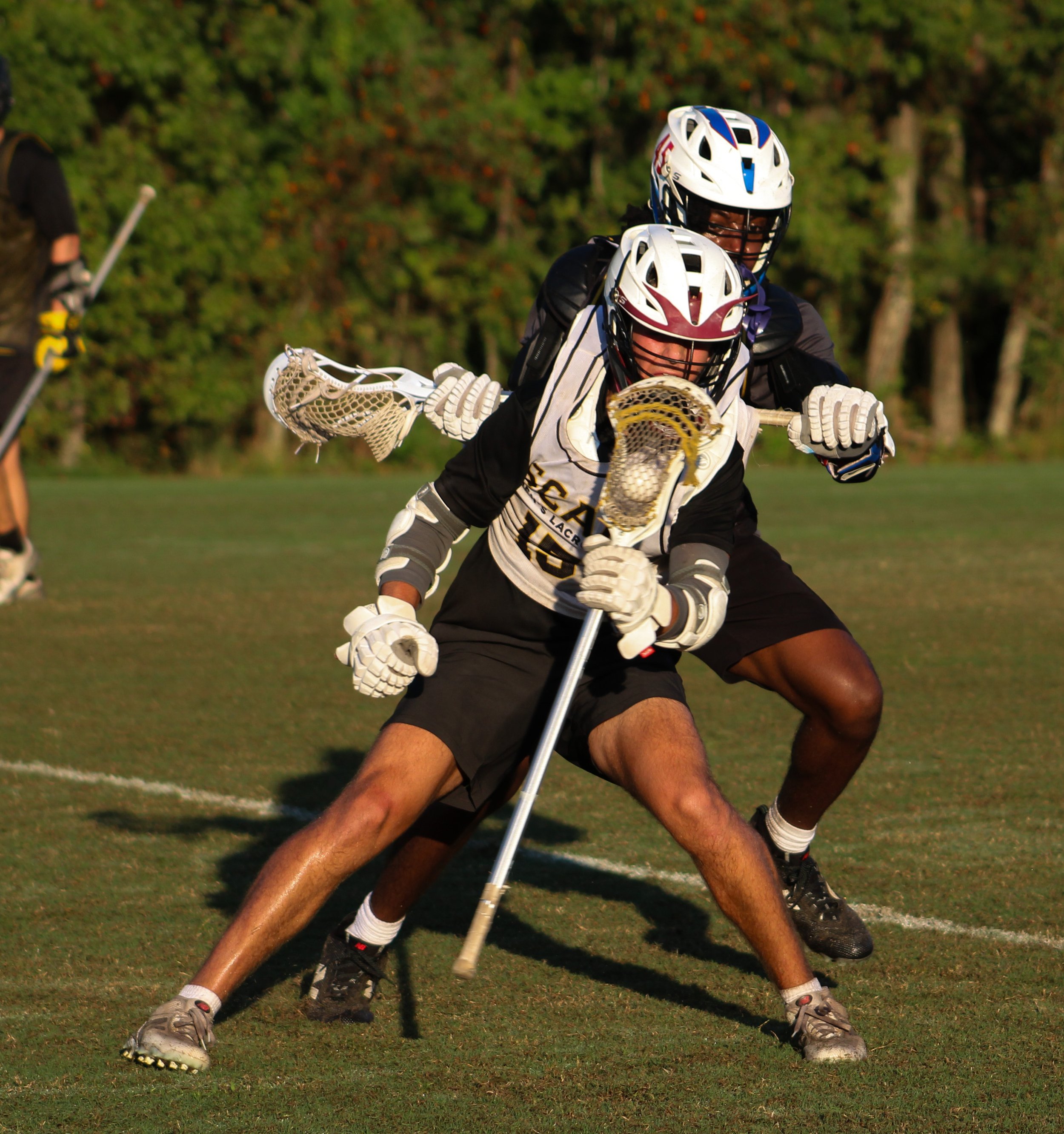 Two lacrosse players in black uniforms play on a grassy field, wearing helmets and gloves, with one player mid-action with a stick and ball, while a defender stands behind.