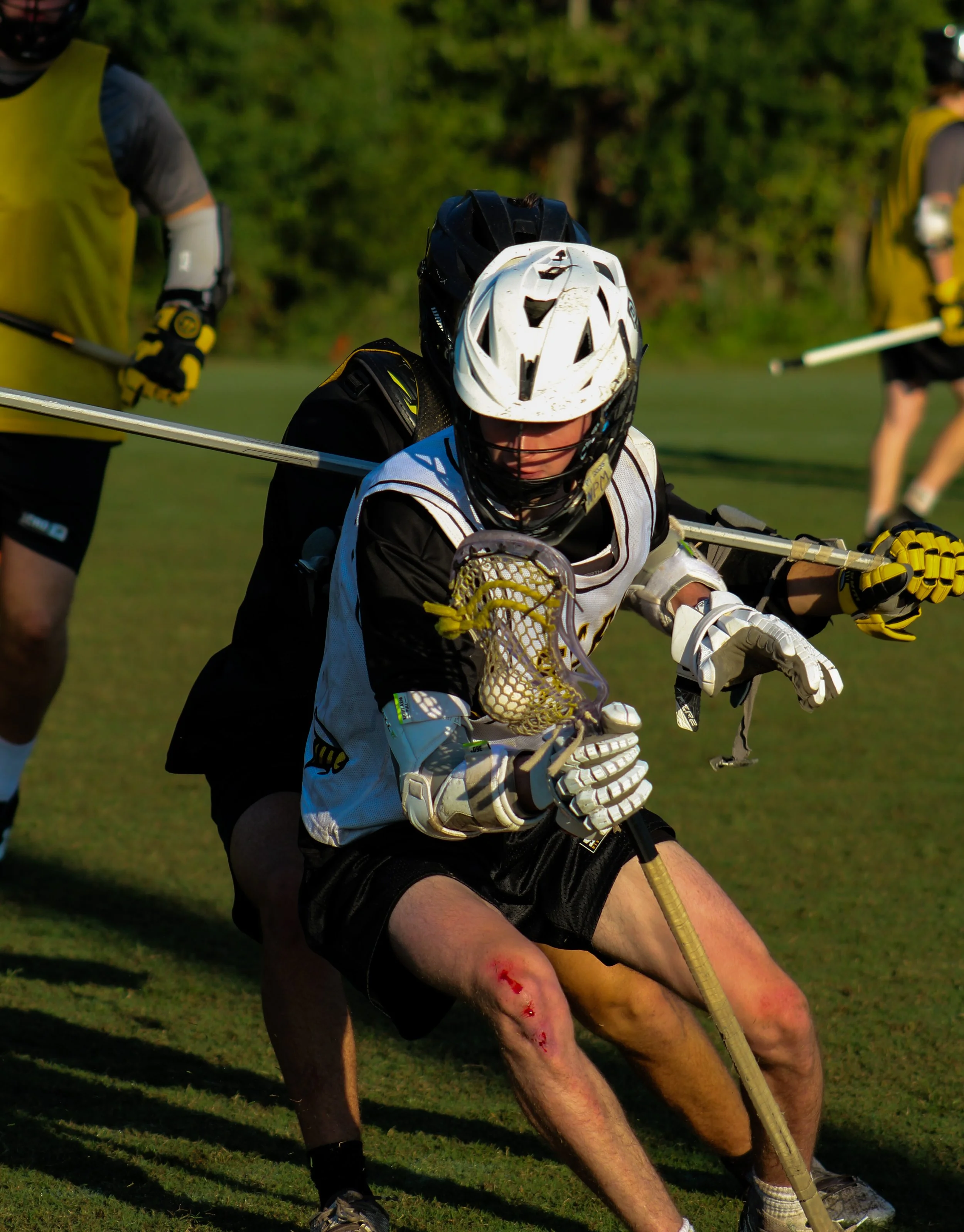 Lacrosse goalie crouching on the field with other players in the background, wearing a white helmet, protective gear, and holding a lacrosse stick with a netted pocket.
