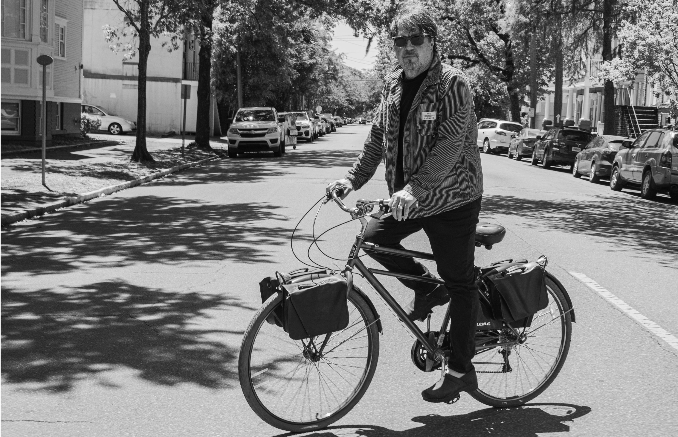 A man with sunglasses riding a bicycle on a quiet street, with parked cars and trees lining the road.