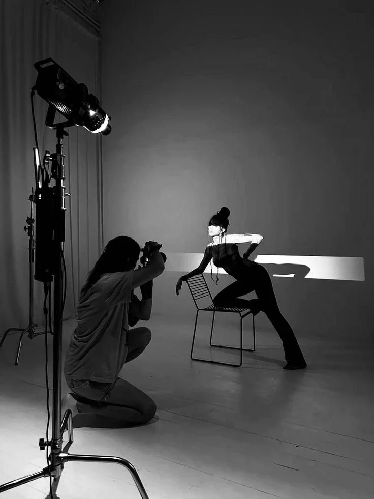 A black and white photo of a photo shoot in a studio with a female model sitting on a chair, styled with a bun hairstyle, and a photographer kneeling on one knee taking pictures. Studio lighting equipment is set up nearby.