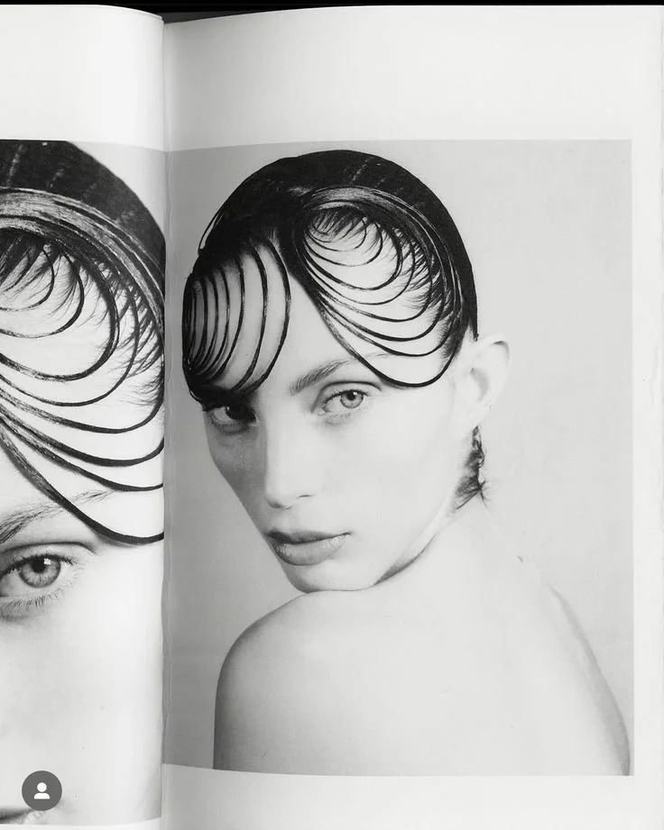 Black and white photograph of a young woman with short, wet-looking hair styled into circular curls, gazing directly at the camera with a serious expression, her shoulder and part of her arm visible.