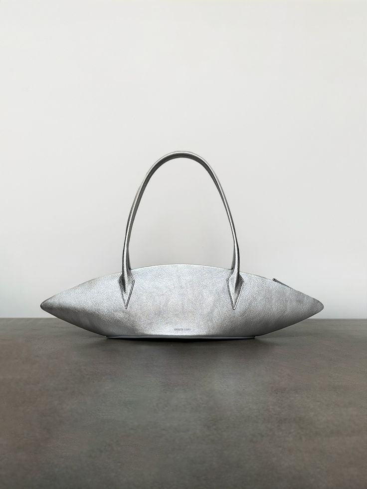 A silver handbag with a unique elongated, rounded shape and double handles, resting on a dark surface with a plain white background.