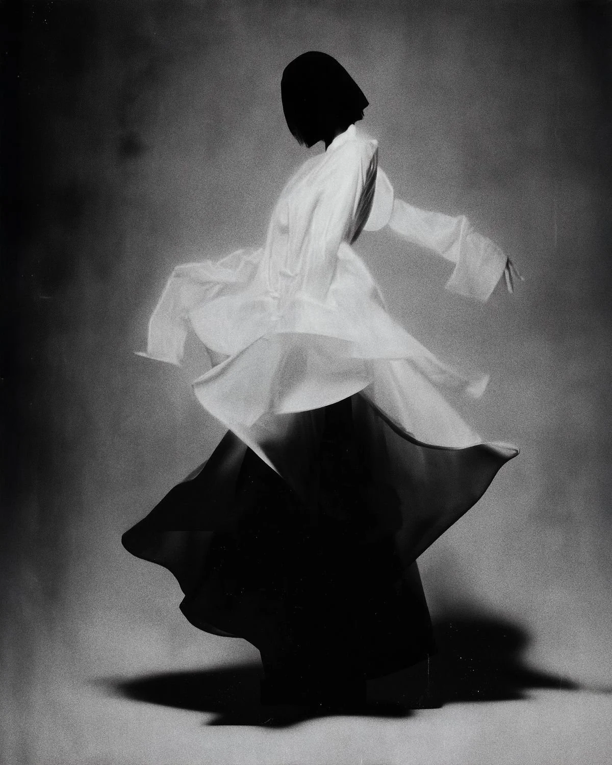 Black and white photo of a woman wearing layered, flowing skirts in motion, with a bob haircut, against a plain background.