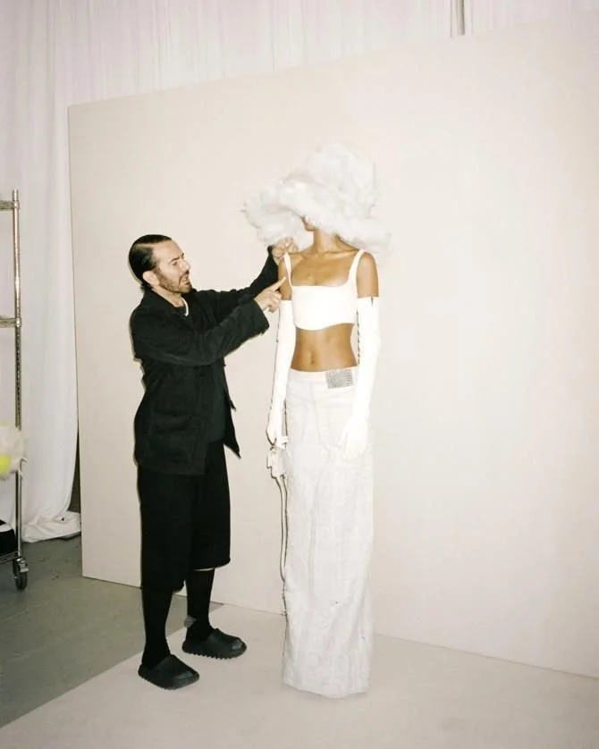 An artist painting a tall, fashionable woman mannequin with a fluffy white wig, white crop top, high-waisted white skirt, and long white gloves in an art studio.