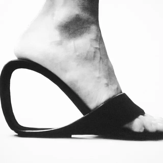 A black and white close-up of a person's foot wearing a high-heeled sandal, with the heel positioned horizontally on the ground.