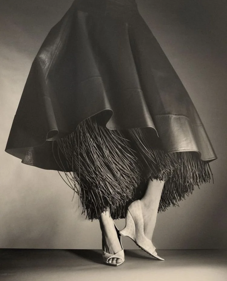 A woman wearing high heels and a voluminous black skirt, with her legs crossed and lifting the skirt slightly to reveal the underside of her shoes and legs.