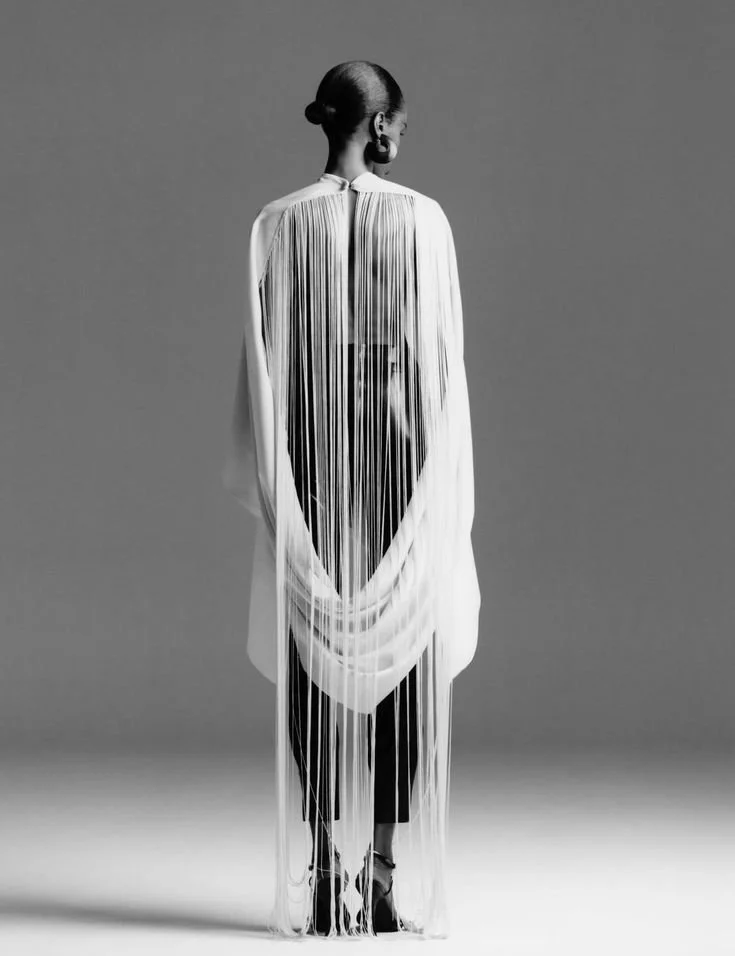 Black and white photo of a woman facing away, wearing a long, semi-transparent dress with vertical stripes, revealing her back and the outline of her back and waist.
