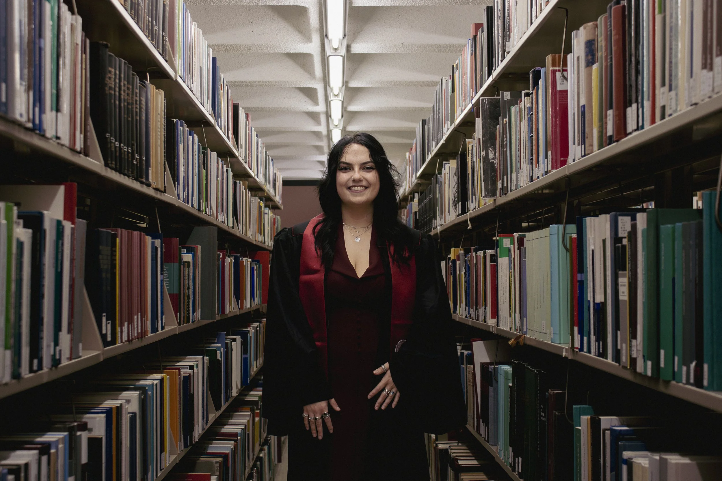 A graduate stands in the middle of an aisle of books in a library. With deeper, moodier, more dramatic lighting, this grad photo session truly stood out. 