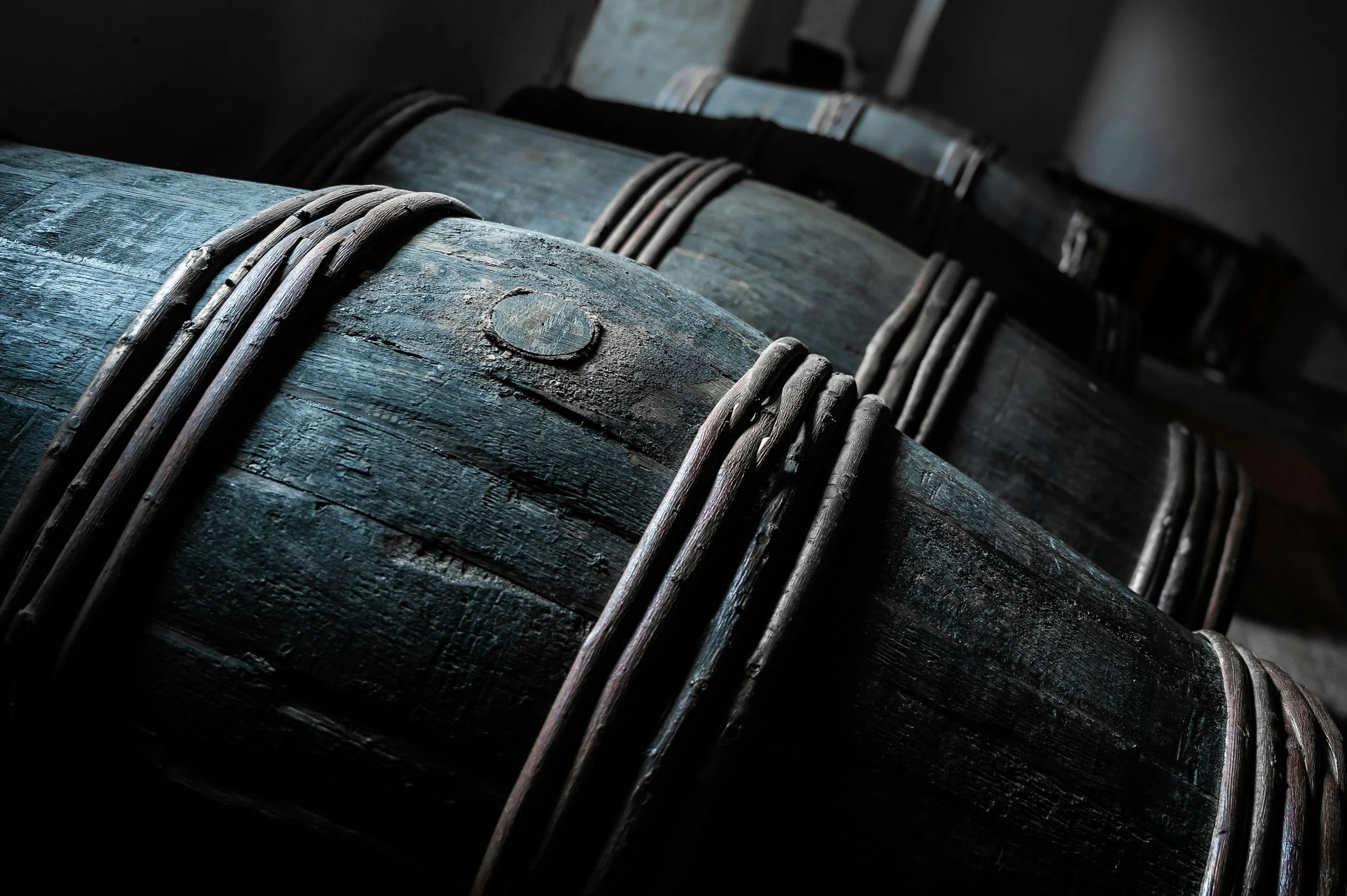 Close-up of dark wooden barrels with metal hoops, stacked in a dimly lit environment, likely a cellar or storage room.