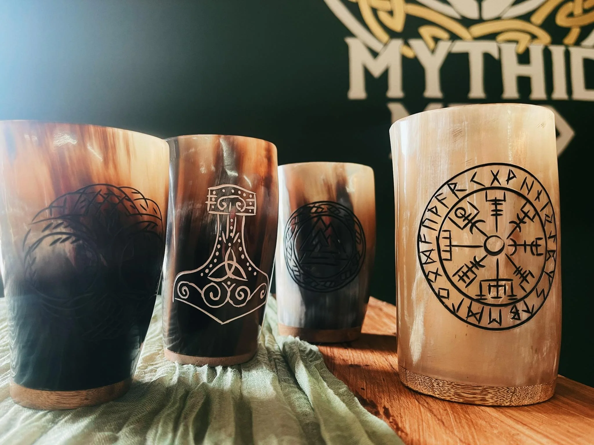 Four cylindrical wooden cups with engraved symbols sitting on a wooden surface, a cloth, and a dark background with partial text 'MYTHIC'.