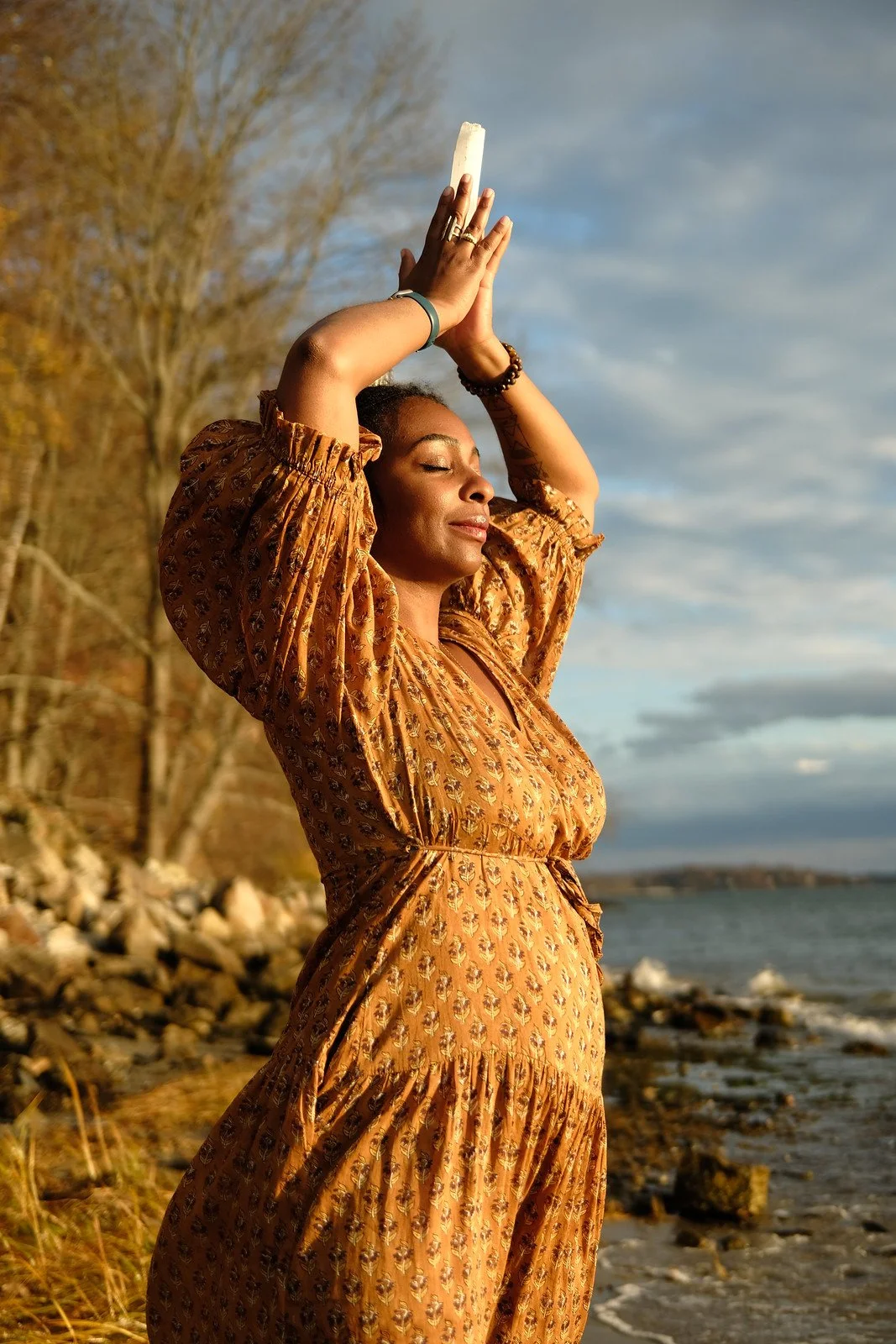 A woman in a patterned orange dress stands on a rocky shoreline with her eyes closed, holding a white object above her head with both hands during sunset, with trees and water in the background.