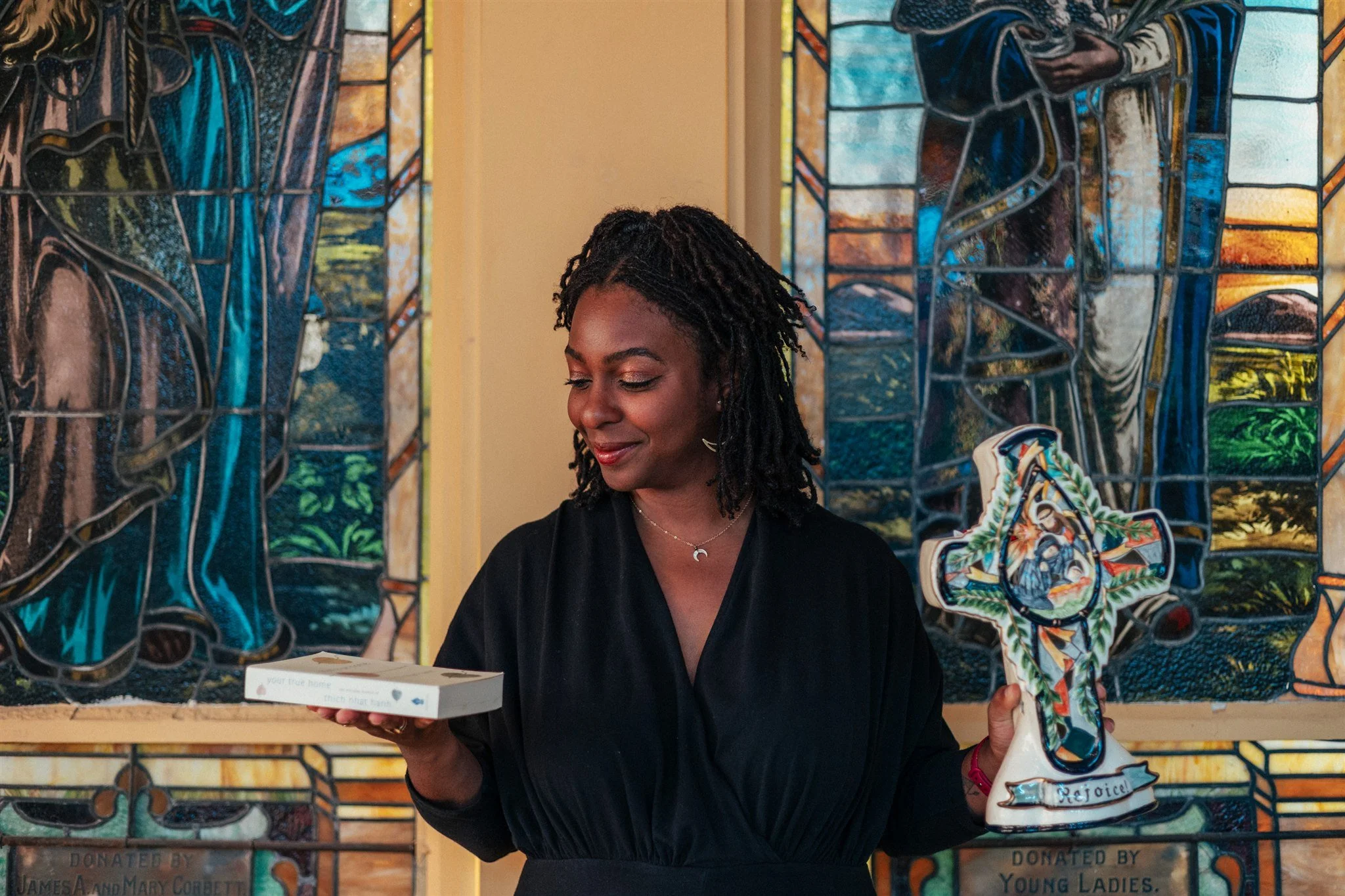 A woman with black curly hair and a black top, smiling and looking at a card, standing in front of stained glass windows and holding a colorful shoe-shaped award.