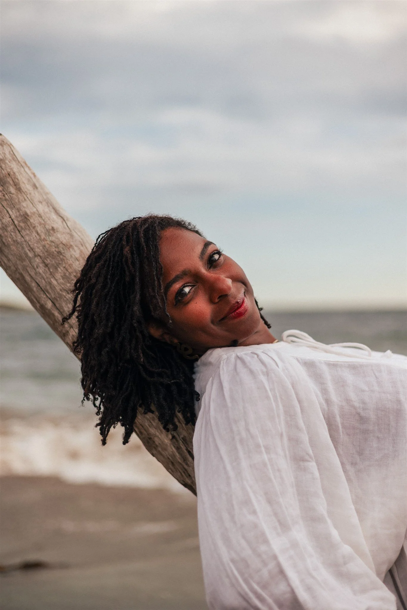 A woman with dark skin and curly hair leaning against a wooden pole on a beach, smiling at the camera with an overcast sky in the background.