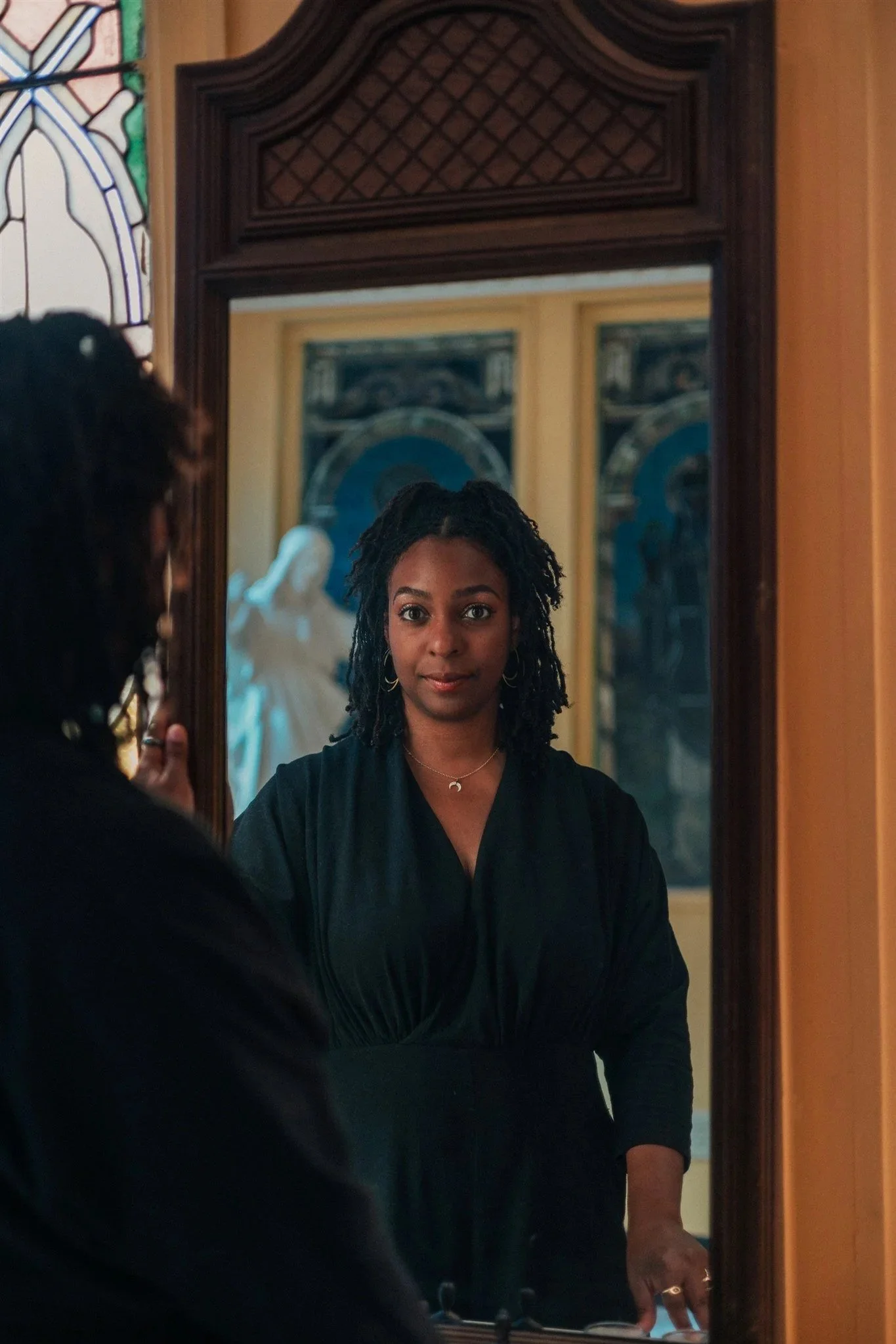 A woman with dark curly hair looking into a decorative wooden framed mirror, with paintings and stained glass window in the background.