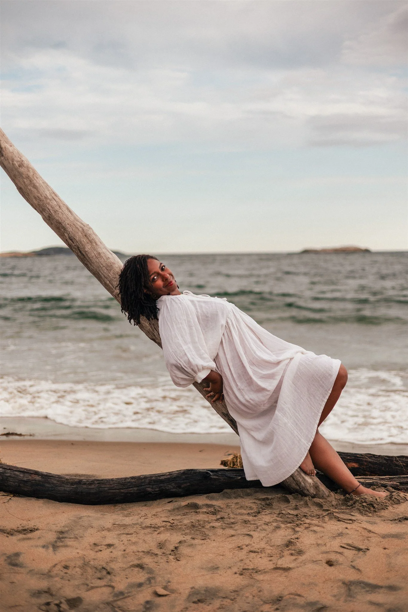 A woman in a white dress leaning against a tilted tree trunk on a beach with ocean in the background during sunset.