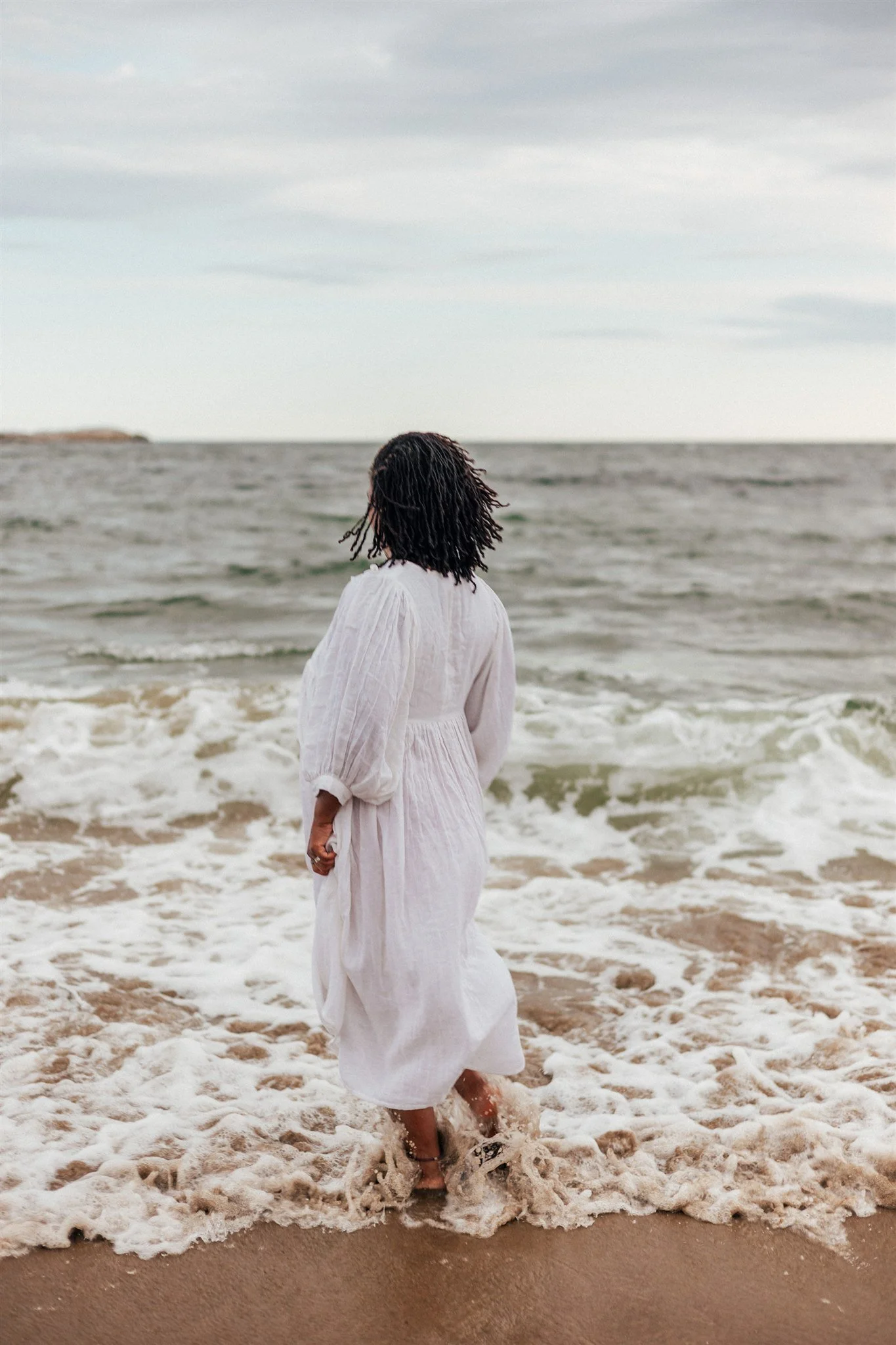 A woman in a white dress walking along the shoreline, with waves approaching her feet at the beach under an overcast sky.