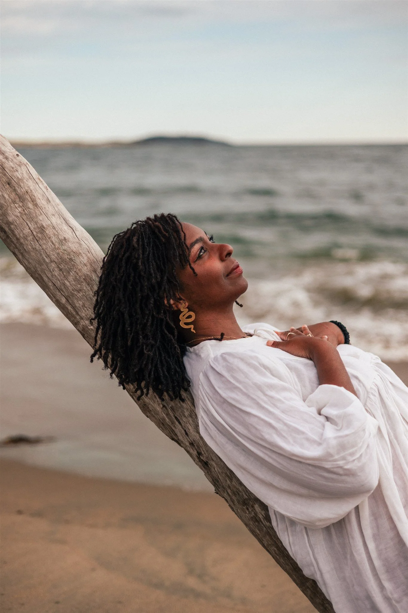 A woman with dreadlocks and gold earrings relaxing on a beach, leaning against a piece of driftwood with her arms crossed, looking up with a peaceful expression at sunset.
