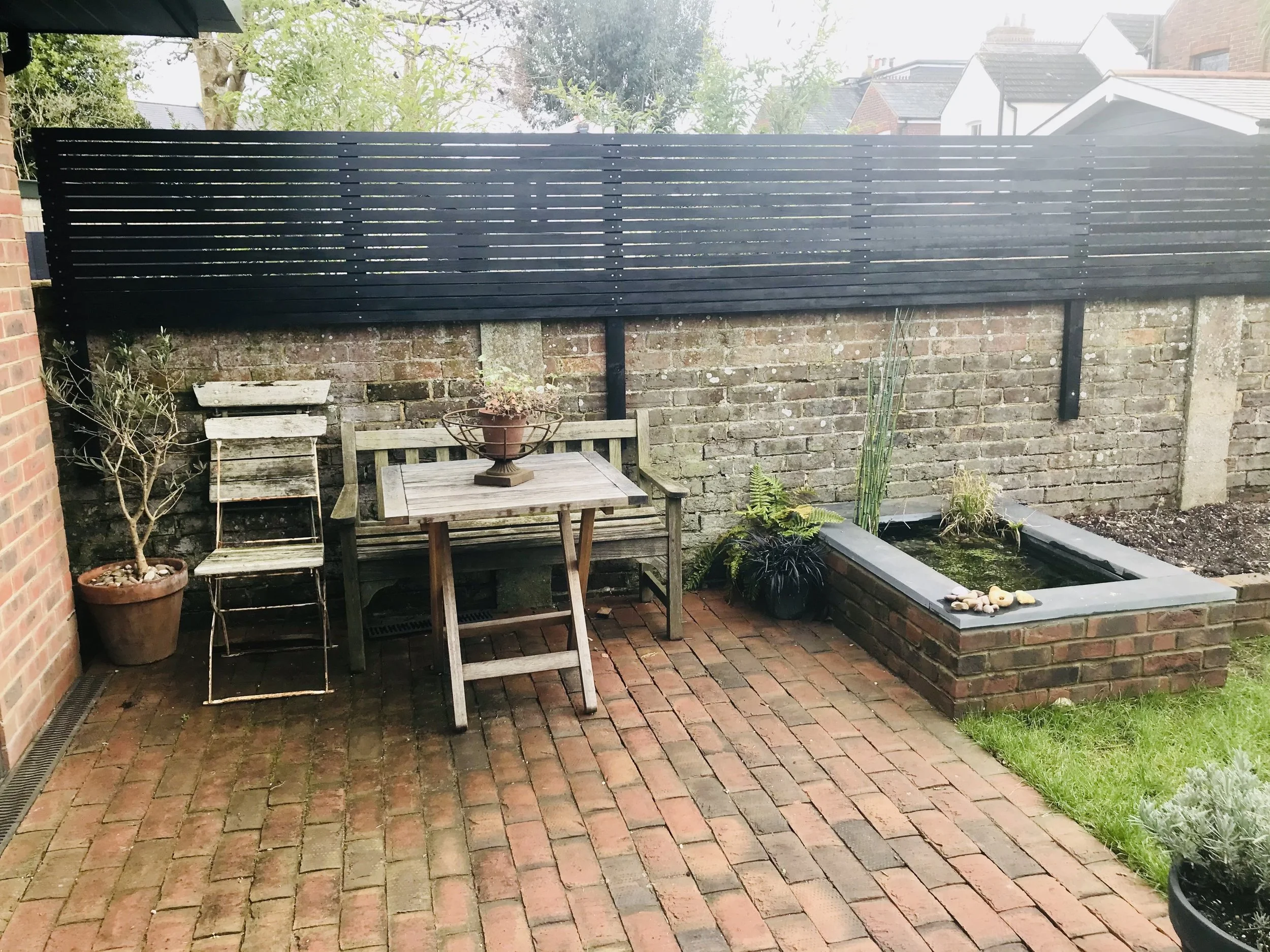 Rustic outdoor patio with brick flooring, wooden bench, table, and potted plants, enclosed by a brick wall and black wooden fence.
