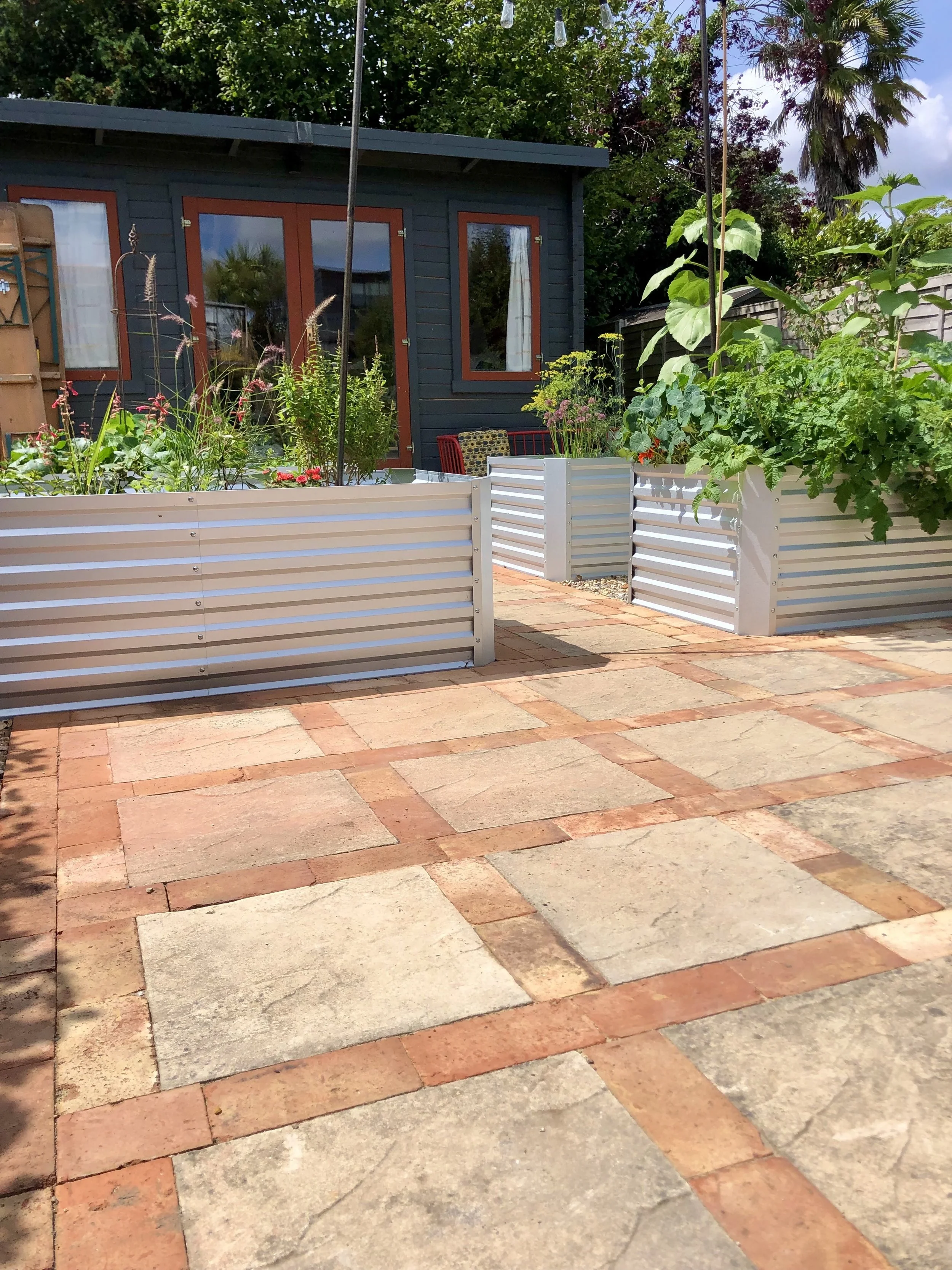 Backyard patio with brick and stone tiles, white raised garden beds filled with plants, and a dark grey house with red-trimmed windows and glass door. Trees and plants surround the area.