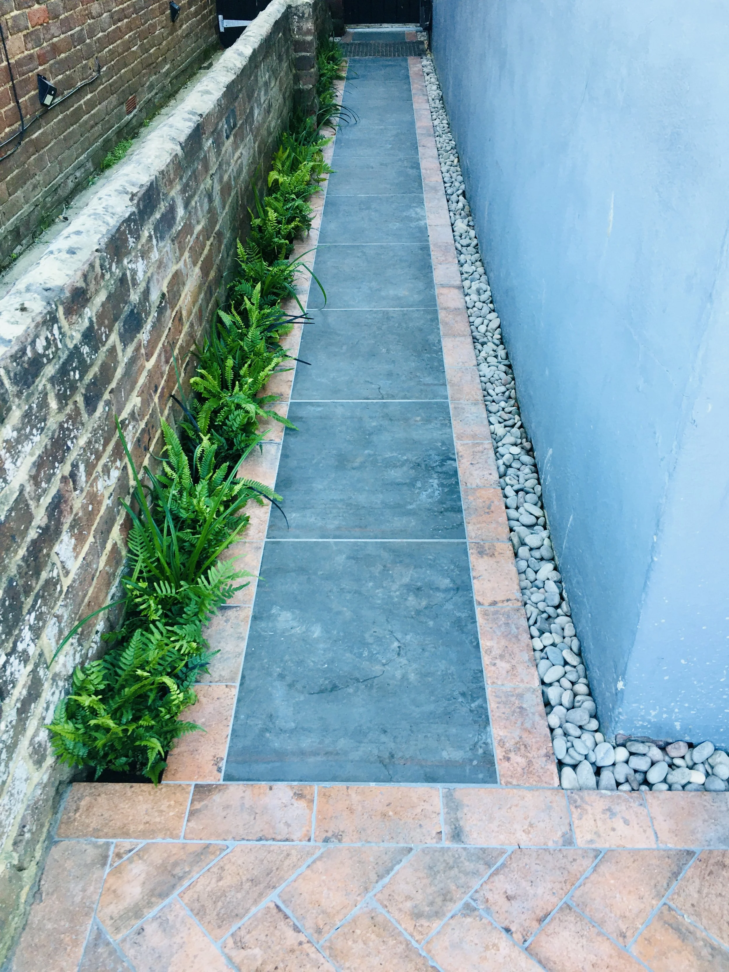 A narrow outdoor pathway with brick and stone borders, a blue wall on the right, a brick wall on the left, green plants along the left side, and smooth white pebbles along the right edge.