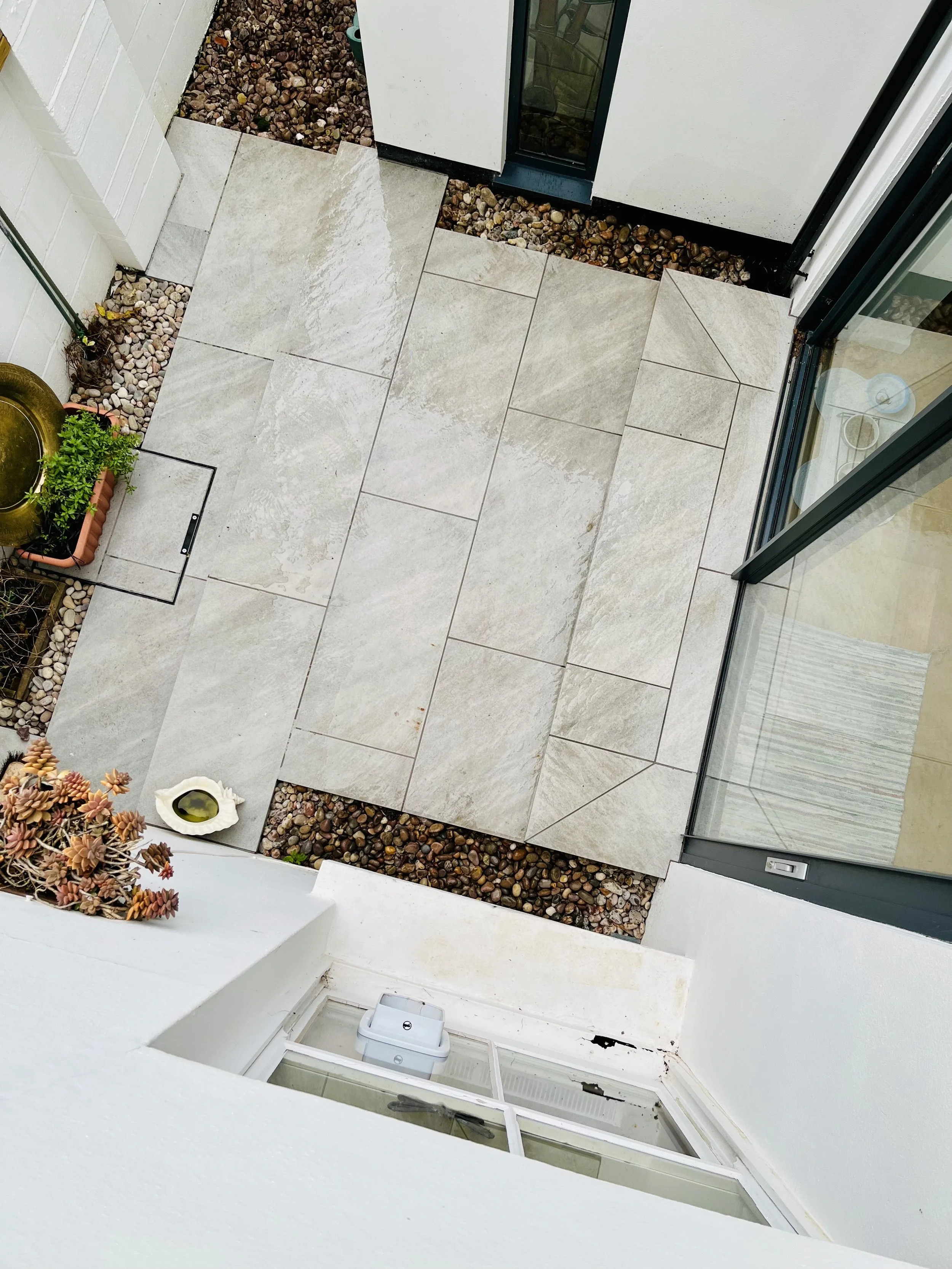 A small outdoor patio with beige tiles surrounded by gravel, a white wall, and a glass door. There is a potted plant, a round bird bath or water feature, and a rectangular hatch on the ground.