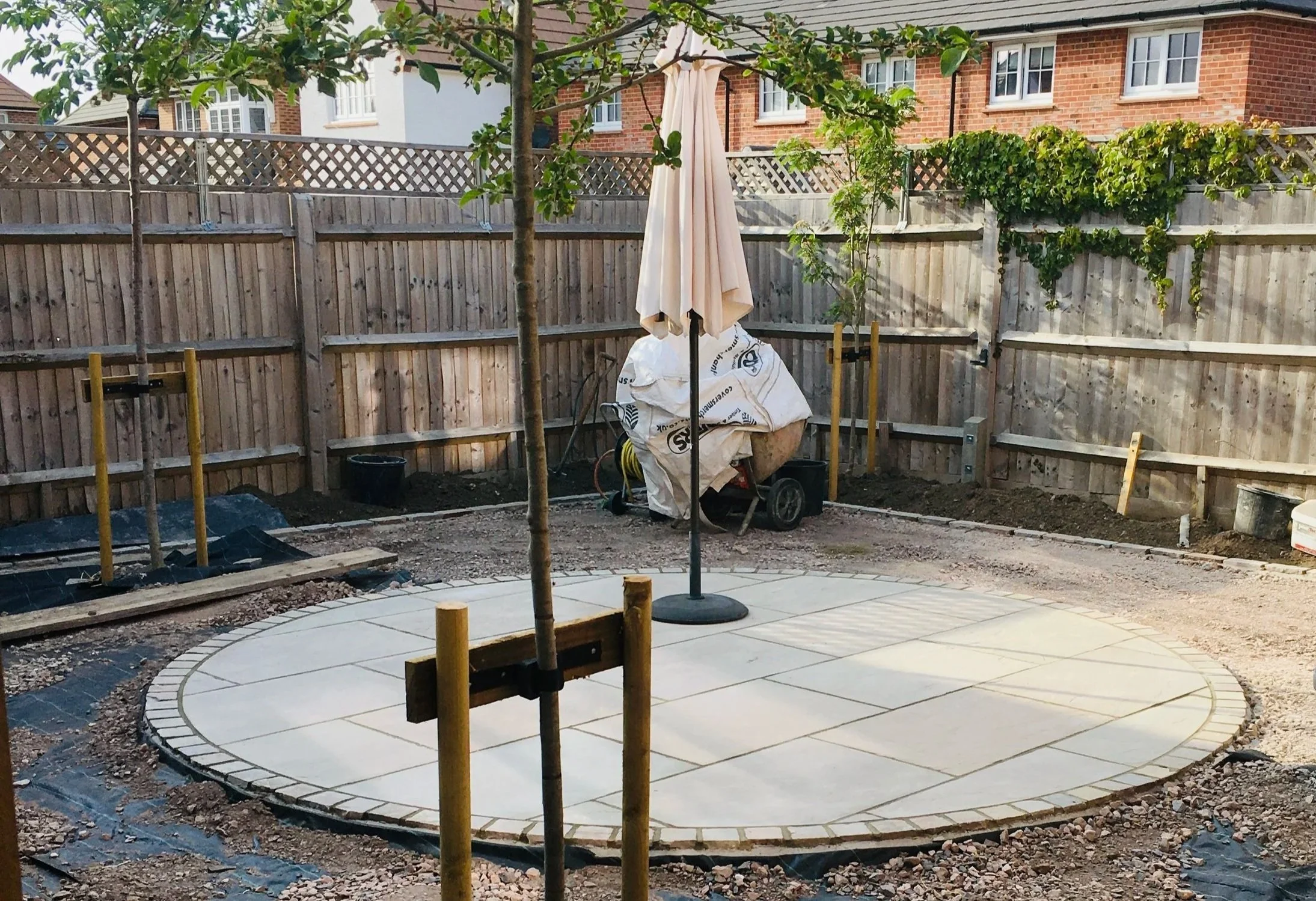 A backyard area under construction with a circular paved patio, a tree in the center, a closed umbrella, and a wooden fence. Construction materials and tools are visible around the site.