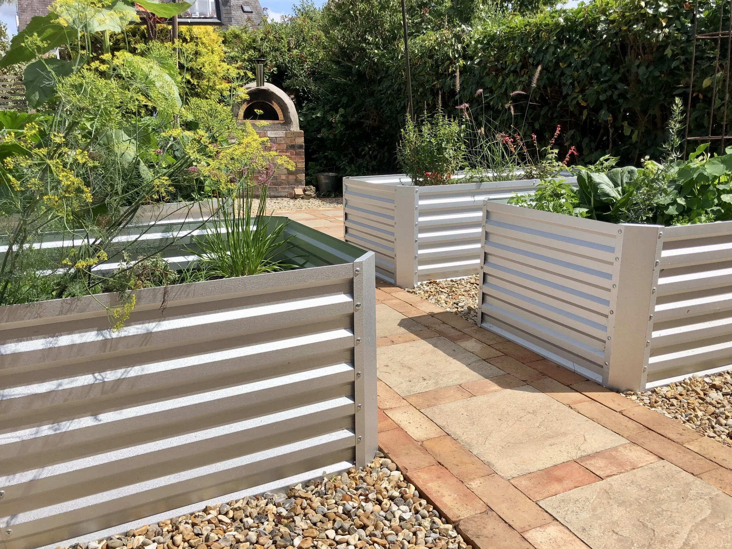 Raised garden beds with various plants in a backyard garden, brick oven in the background, paved pathways, and gravel ground cover.