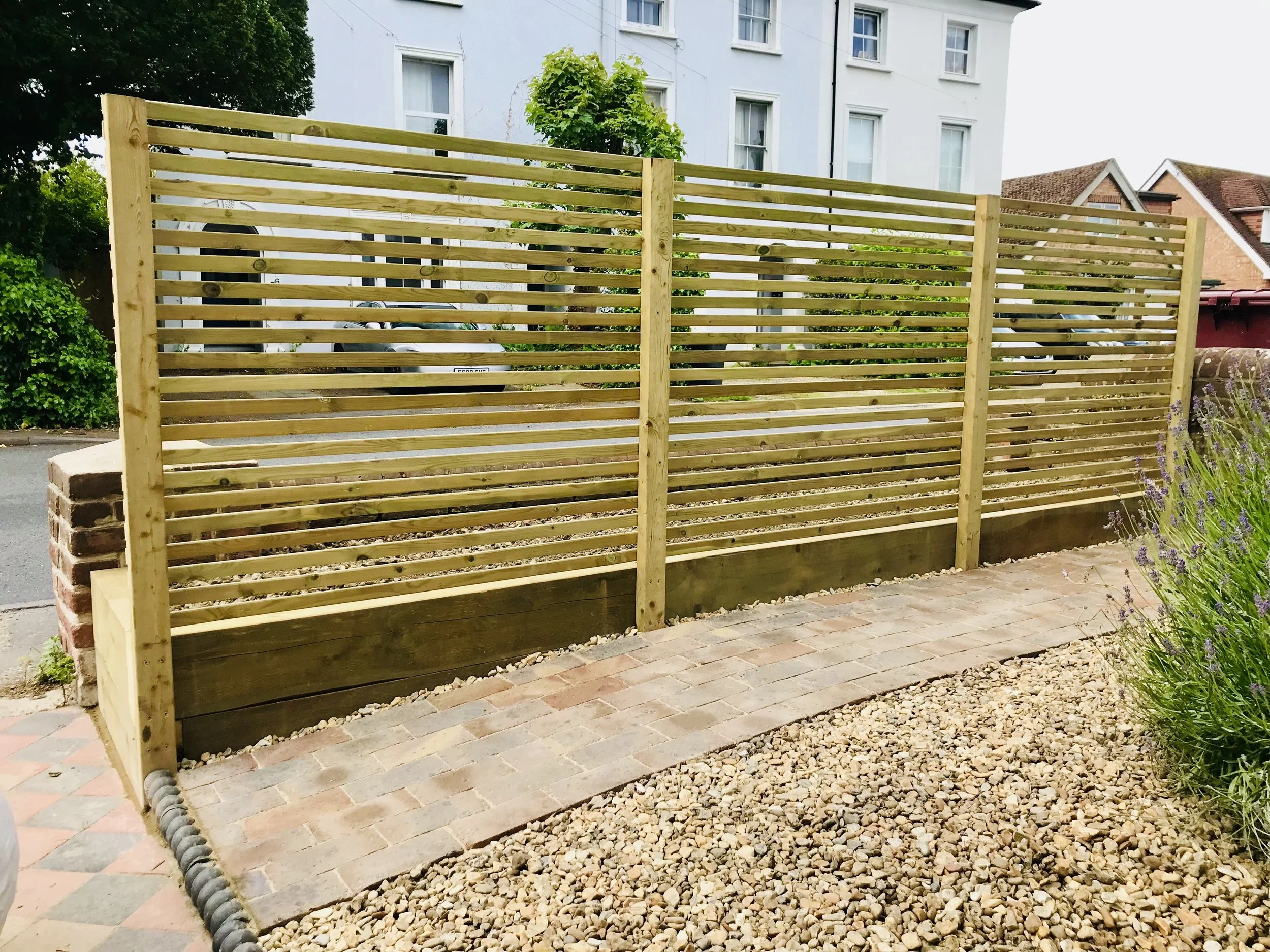 New wooden slat privacy fence installed beside a brick walkway and gravel garden, with residential buildings in the background.